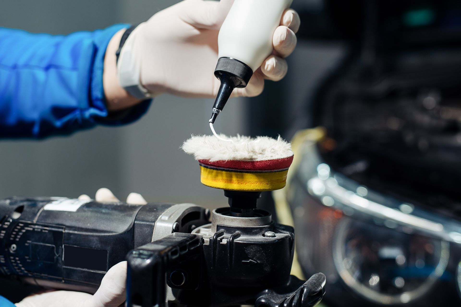 A gloved hand applies polishing compound to a buffer pad on a car, detailer at work.