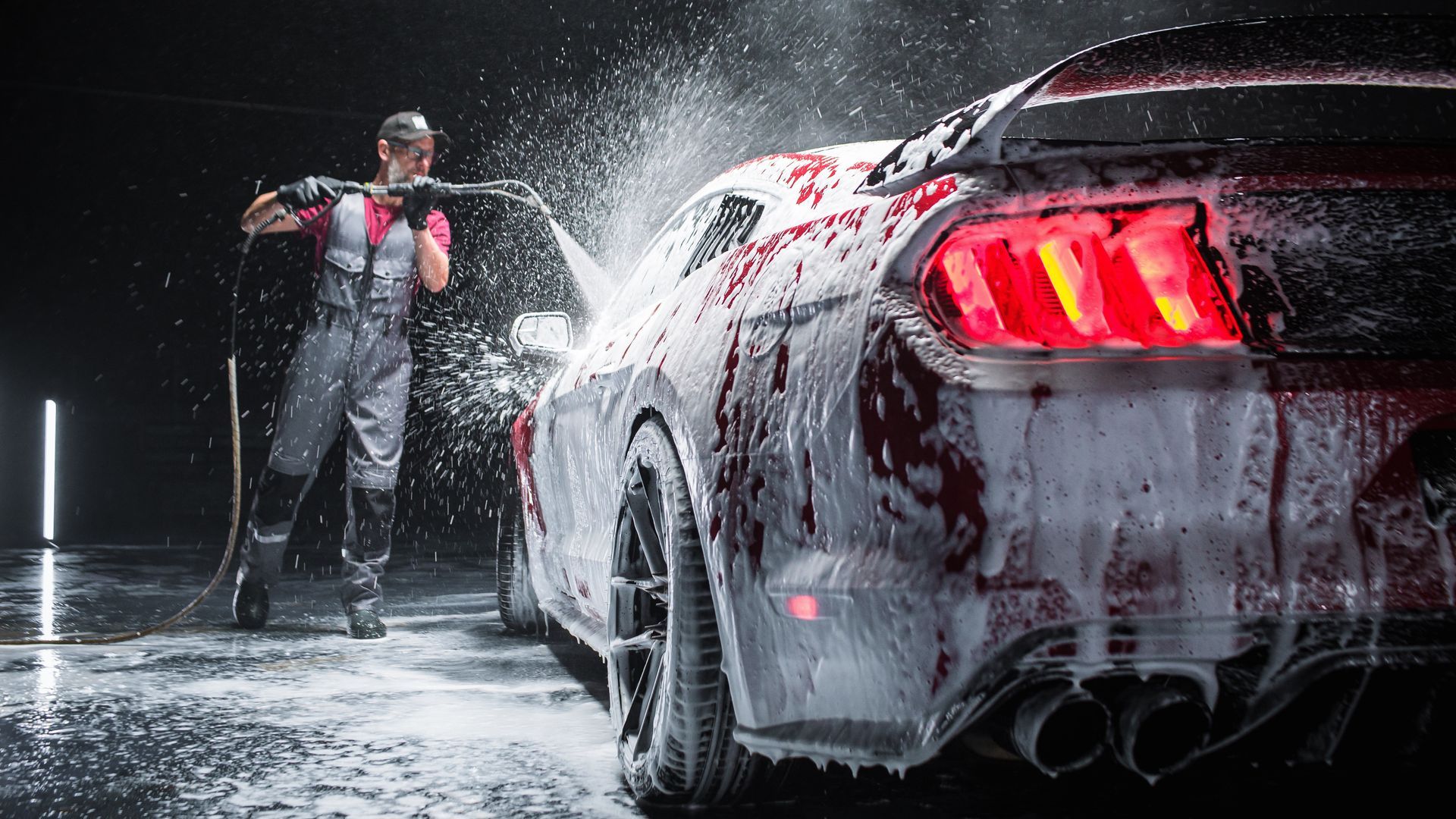 Man washing a red sports car with foam and a power washer in a dark studio.