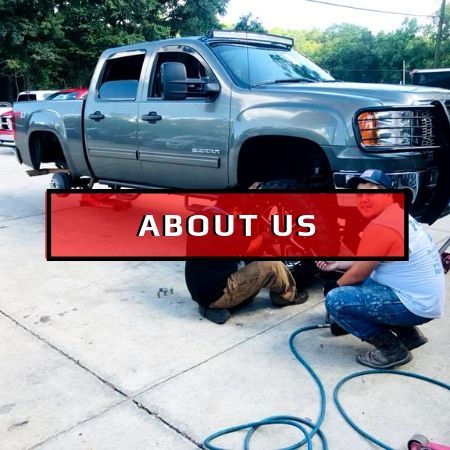 Two people work on a lifted gray pickup truck outdoors, with a red