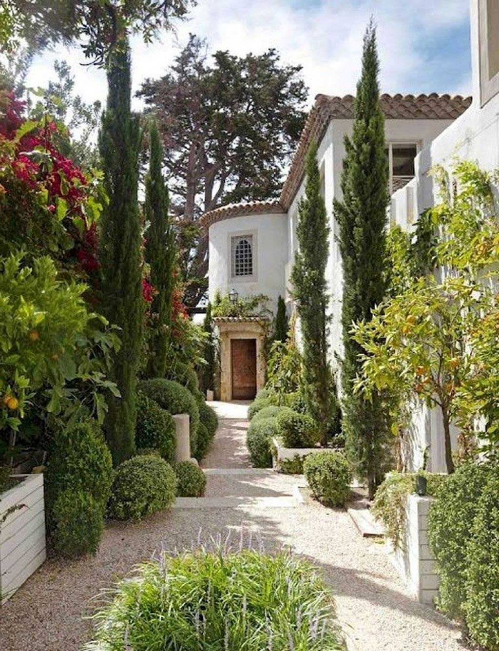 Gravel pathway leading to a white stucco building with a wooden door, flanked by green hedges and tall cypress trees.