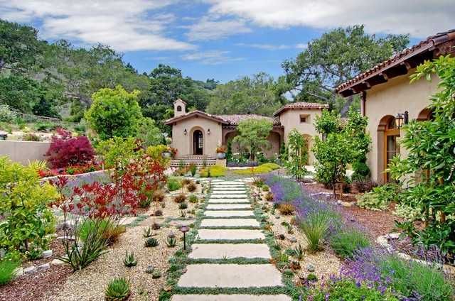 Stone path leads through a colorful garden to a Spanish-style house under a blue sky.
