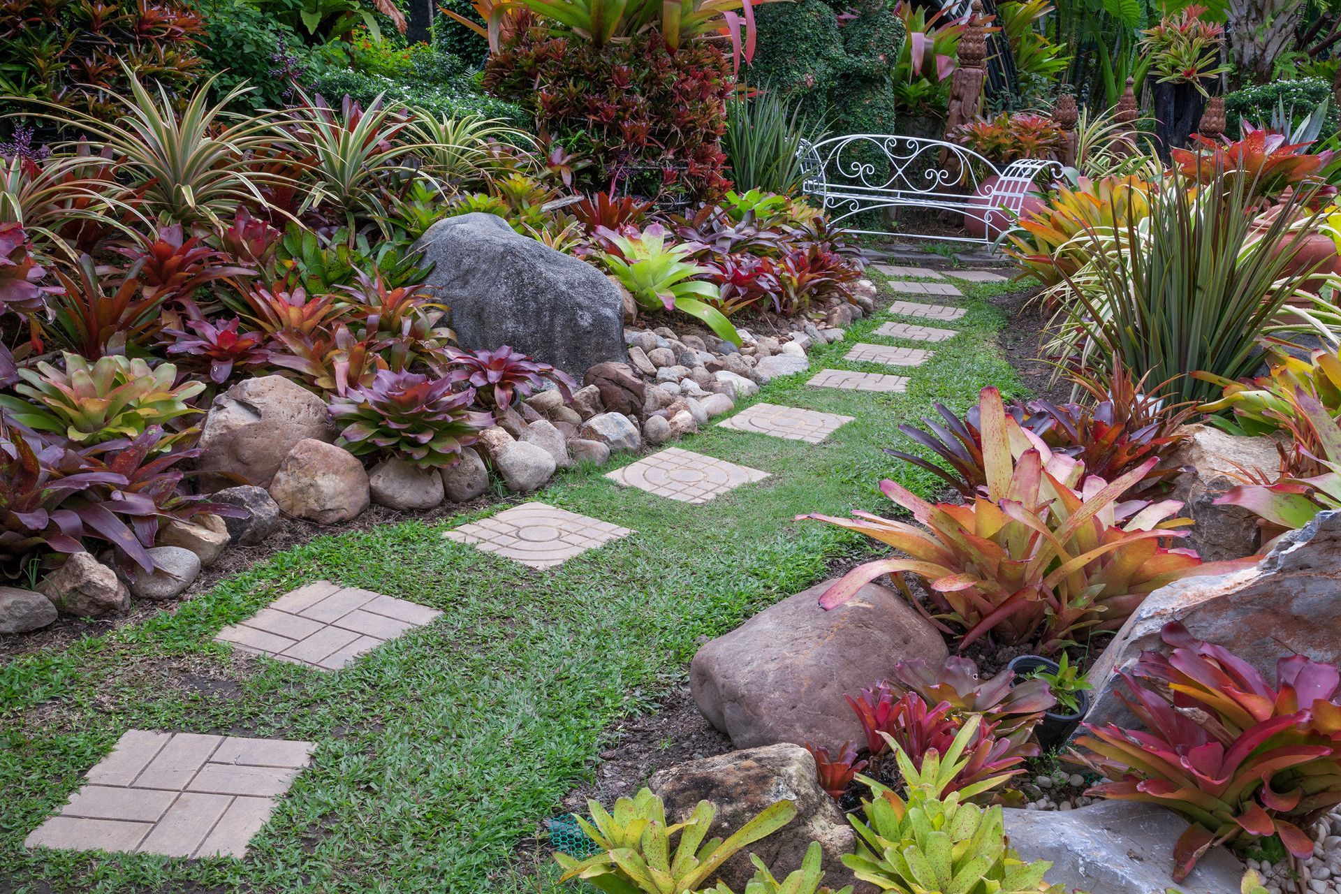 Stone pathway through a vibrant garden with colorful plants, rocks, and a bench in the distance.
