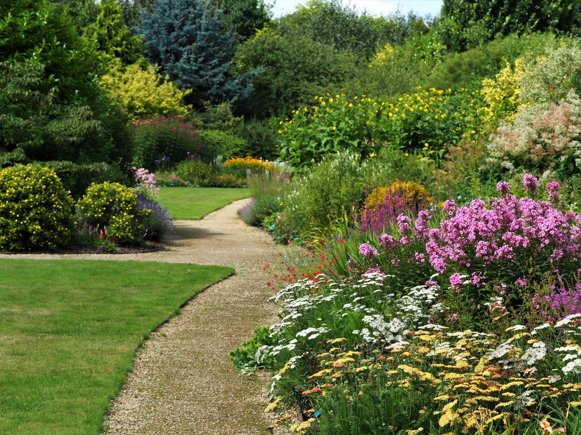 Gravel path winds through a vibrant garden with colorful flowers, green grass, and lush trees.