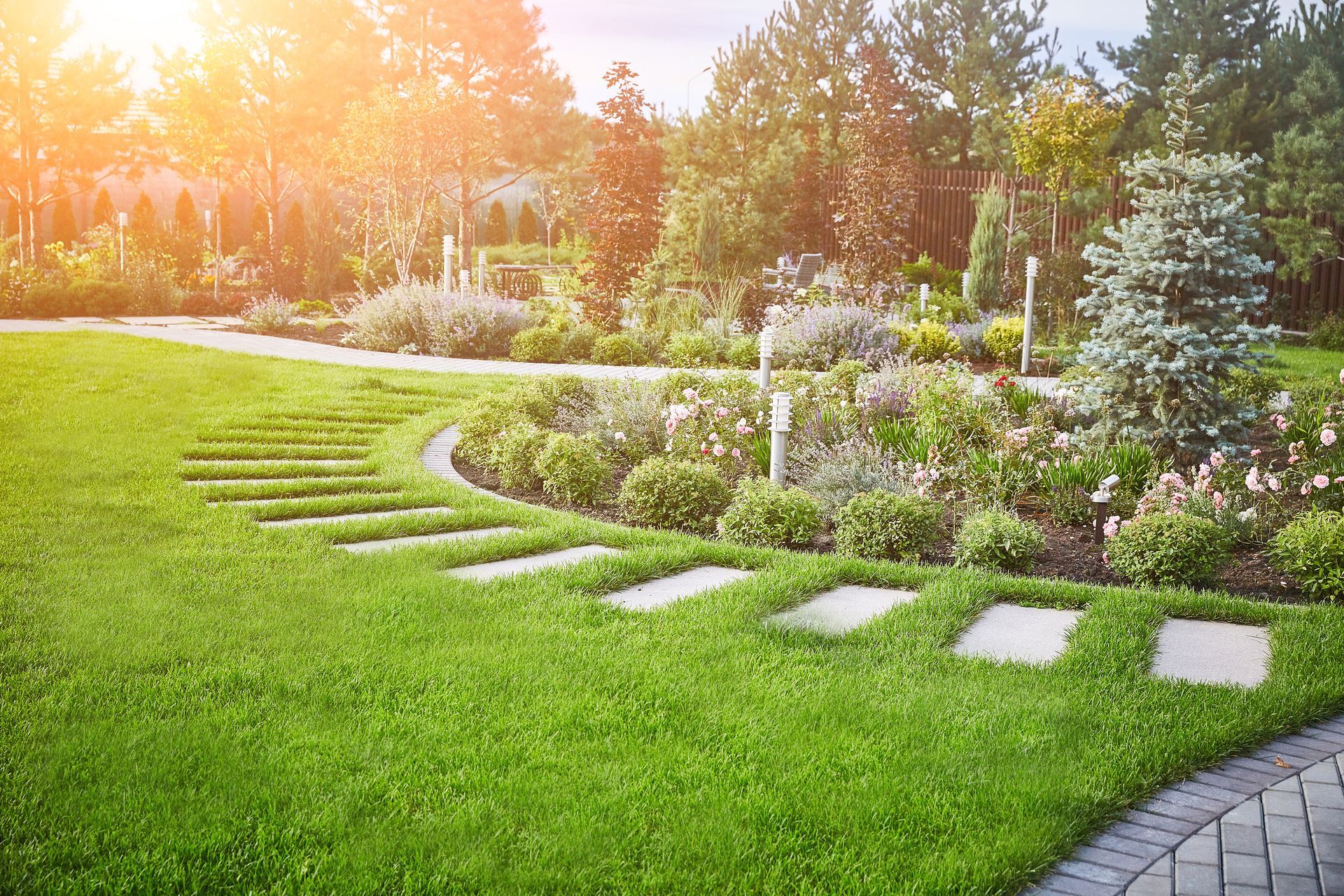Lush green lawn with stone path leading through a vibrant garden, bright sunlight overhead.