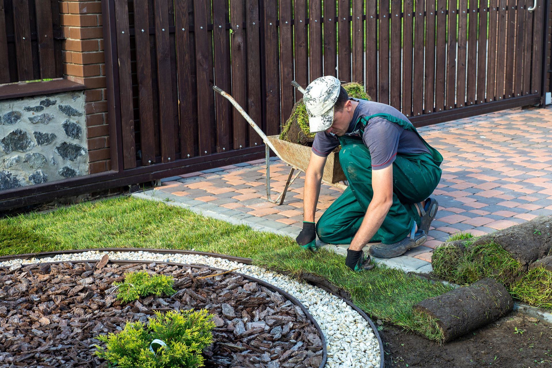 Man laying sod near a garden bed and brick patio. Dark green work clothes, brown fence in background.
