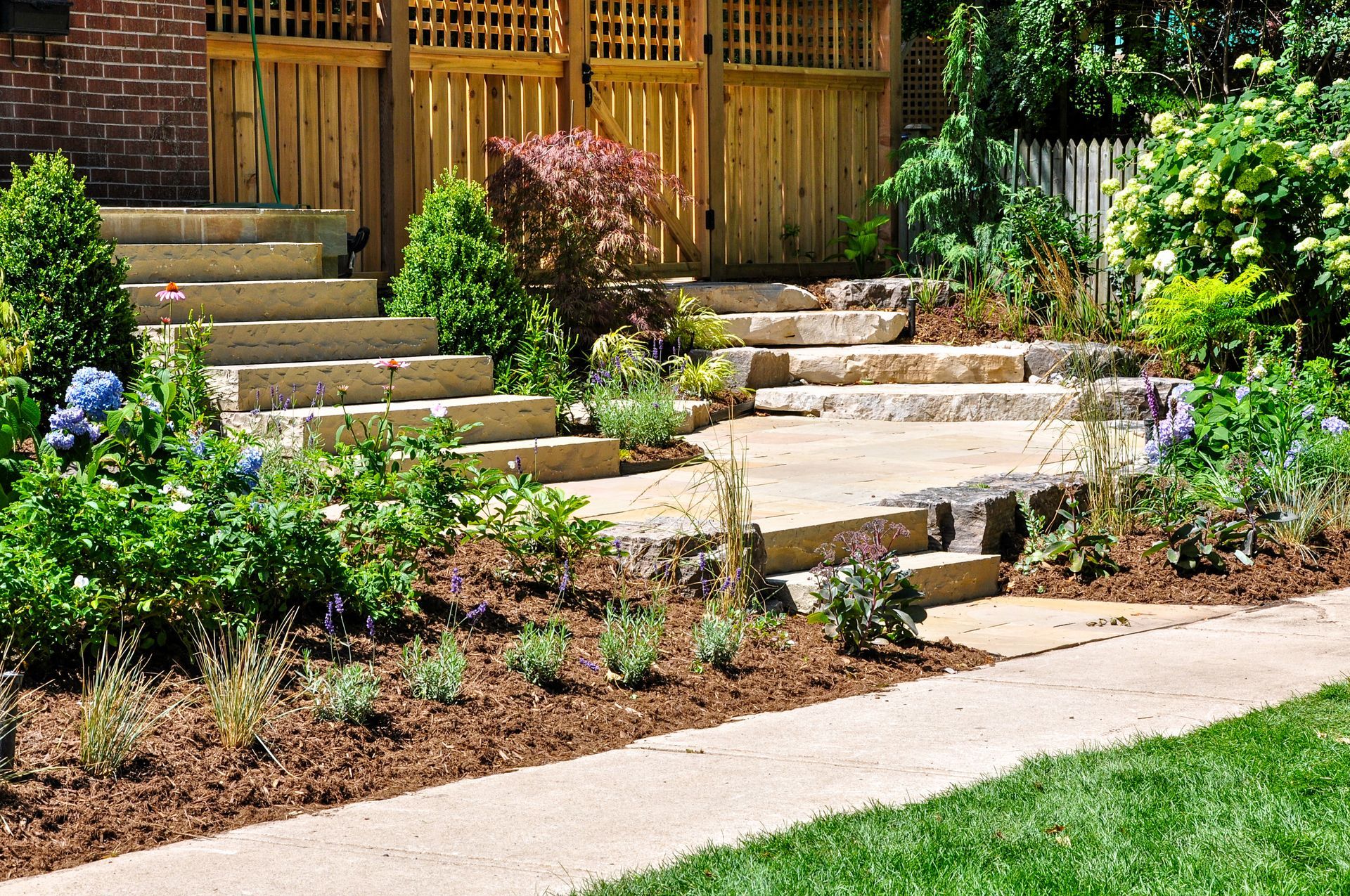 Stone steps leading to a wooden gate, surrounded by garden beds with green plants and mulch, near a concrete path.