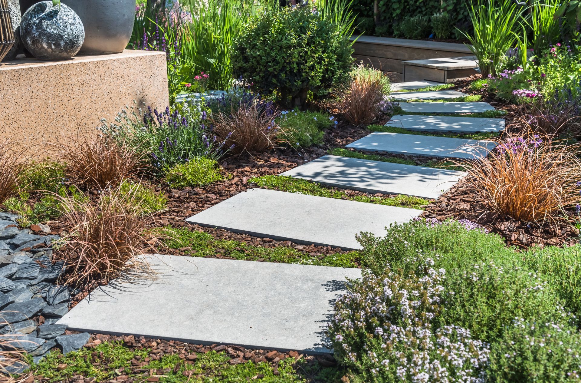 Stepping stones lead through a garden with green plants and decorative bark mulch.
