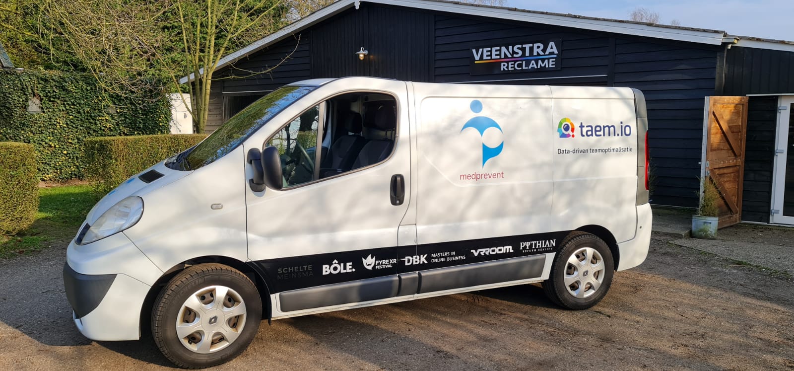 Een wit busje staat geparkeerd voor een zwart gebouw.