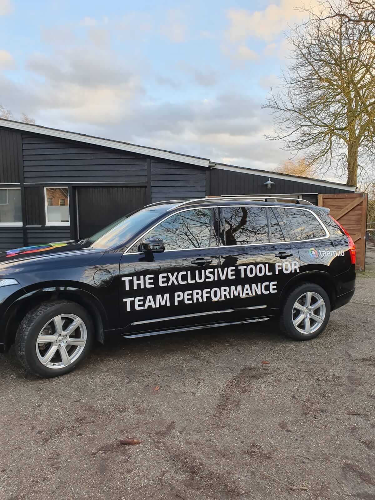 Een zwarte SUV met op de zijkant de tekst 'Exclusieve tool voor teamprestaties' staat geparkeerd voor een gebouw.