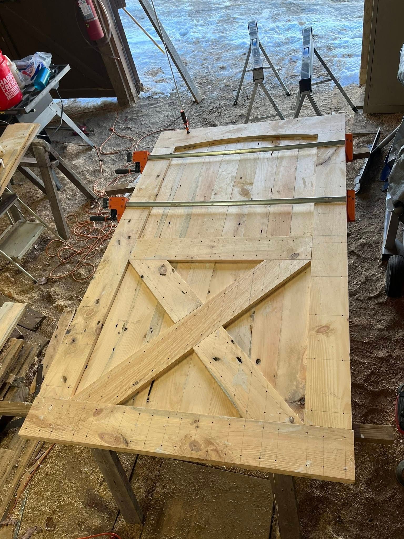 A wooden barn door under construction lies on sawhorses in a workshop, featuring a cross-brace design and visible clamps.