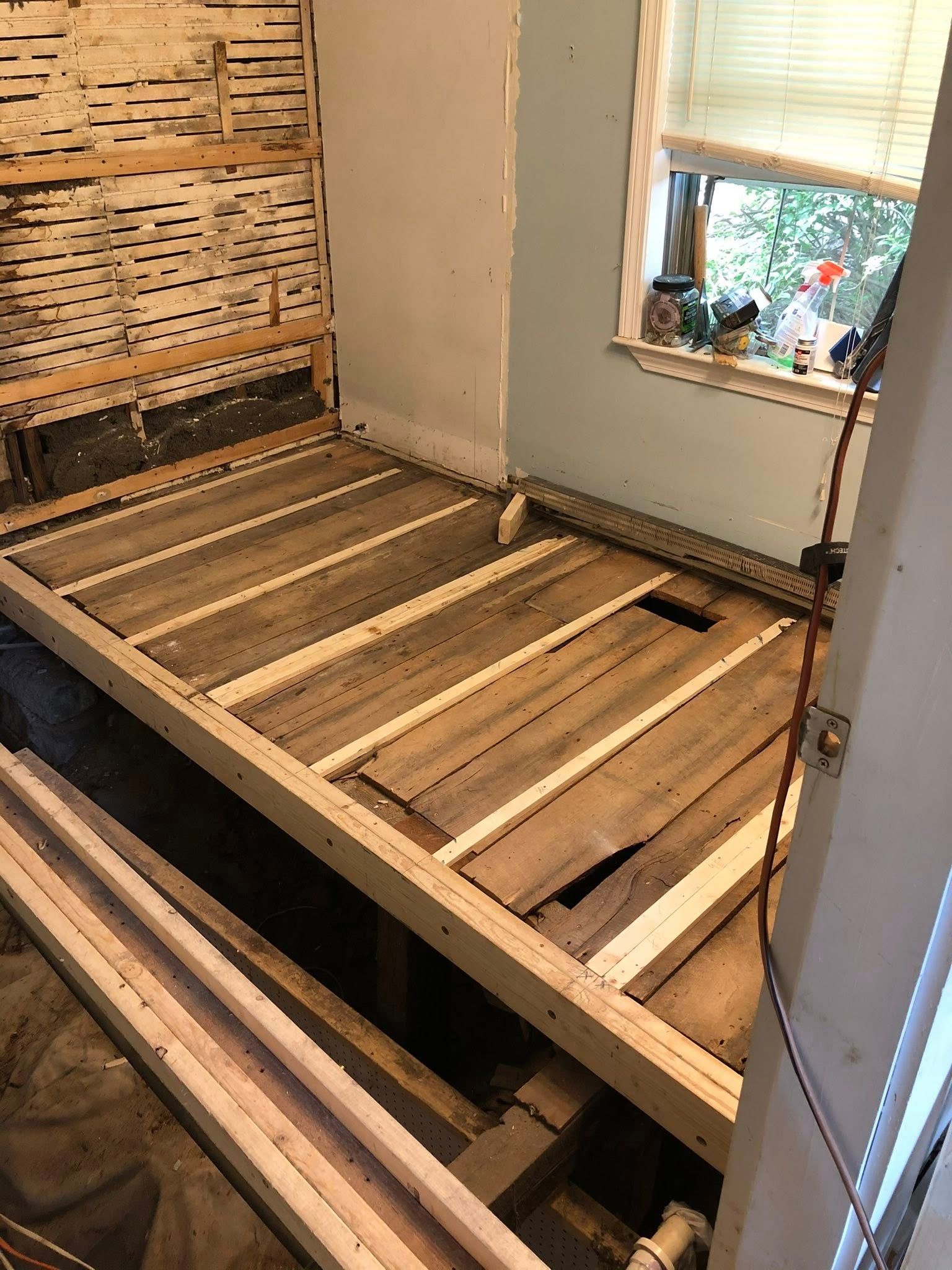 A room under renovation with stripped wall lath and exposed subflooring on the joists, next to a window.