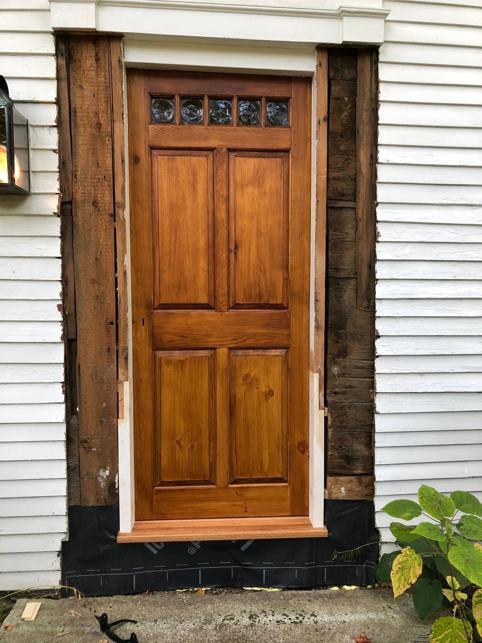 A wooden door in a white house, undergoing a renovation with missing exterior trim exposing the rough frame and wall.