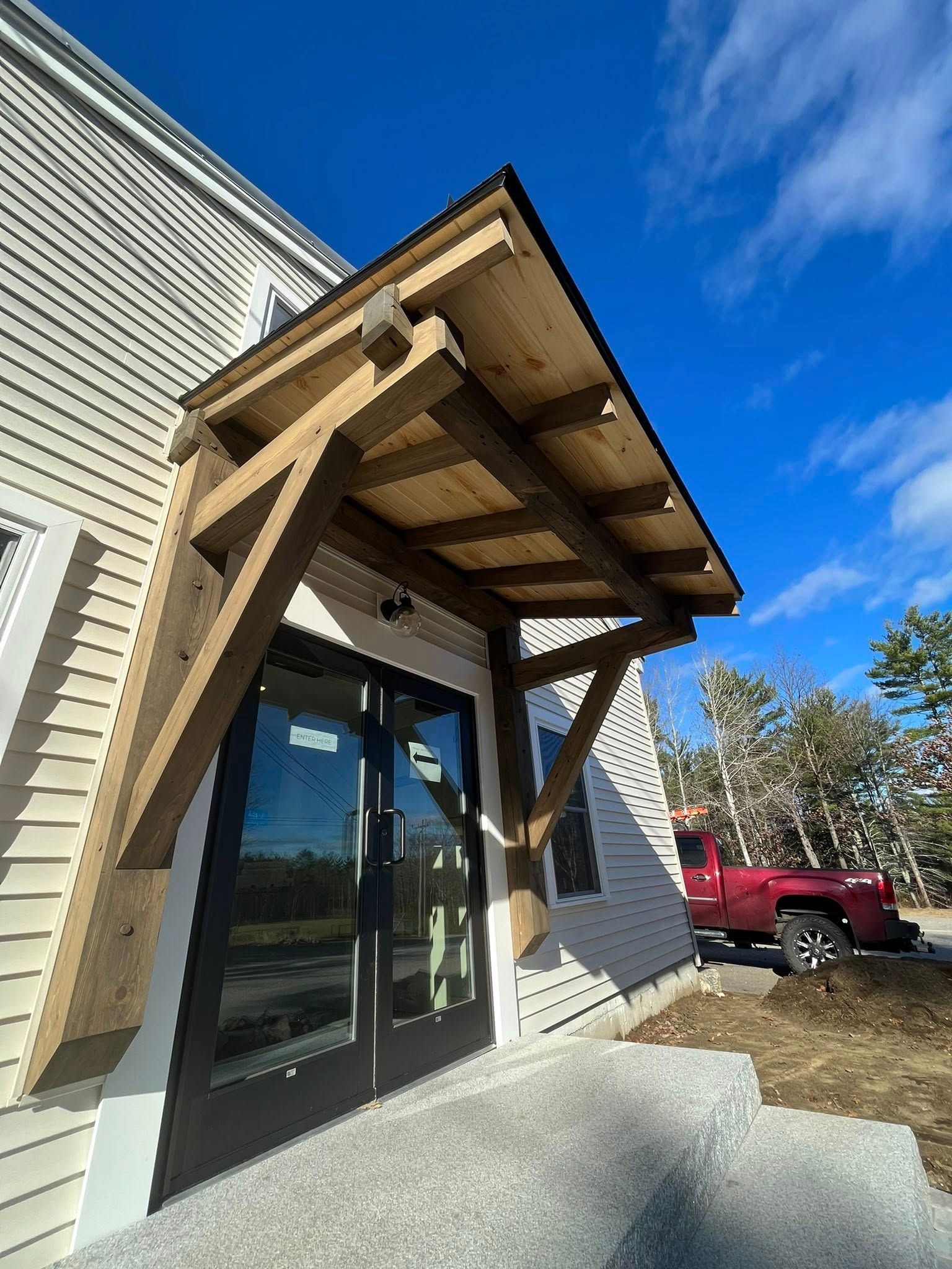 A wooden porch overhang above a double glass door on the side of a light-colored building with a red truck nearby.