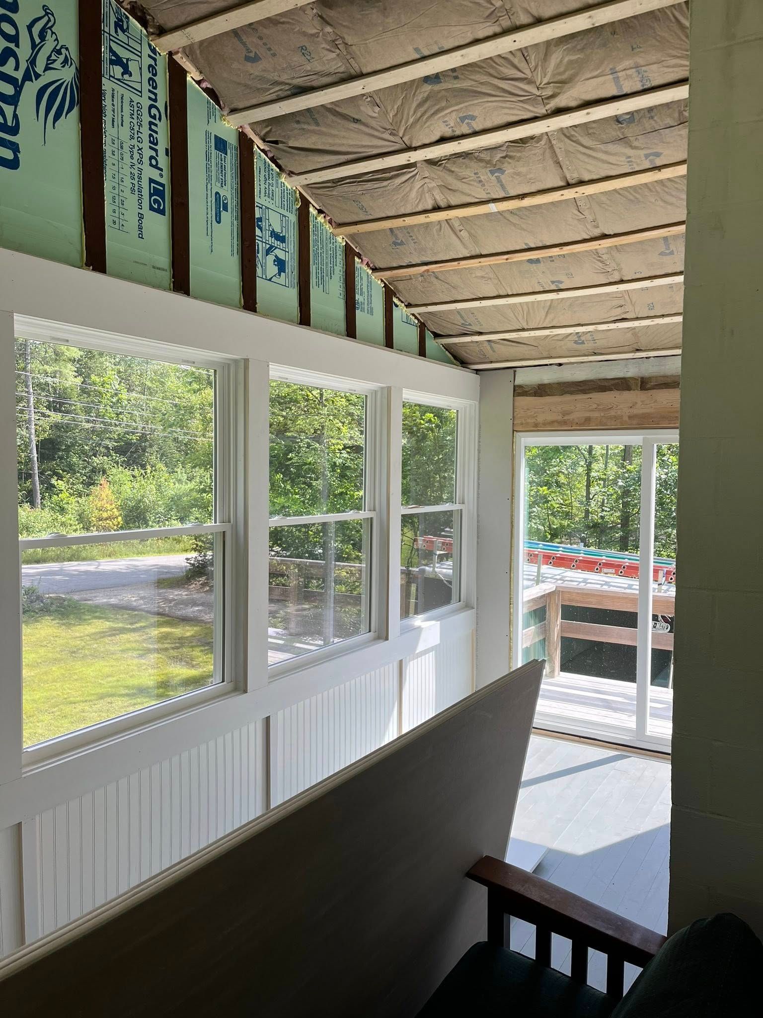 A room under construction with white-framed windows, exposed ceiling joists, and green insulation panels on the walls.