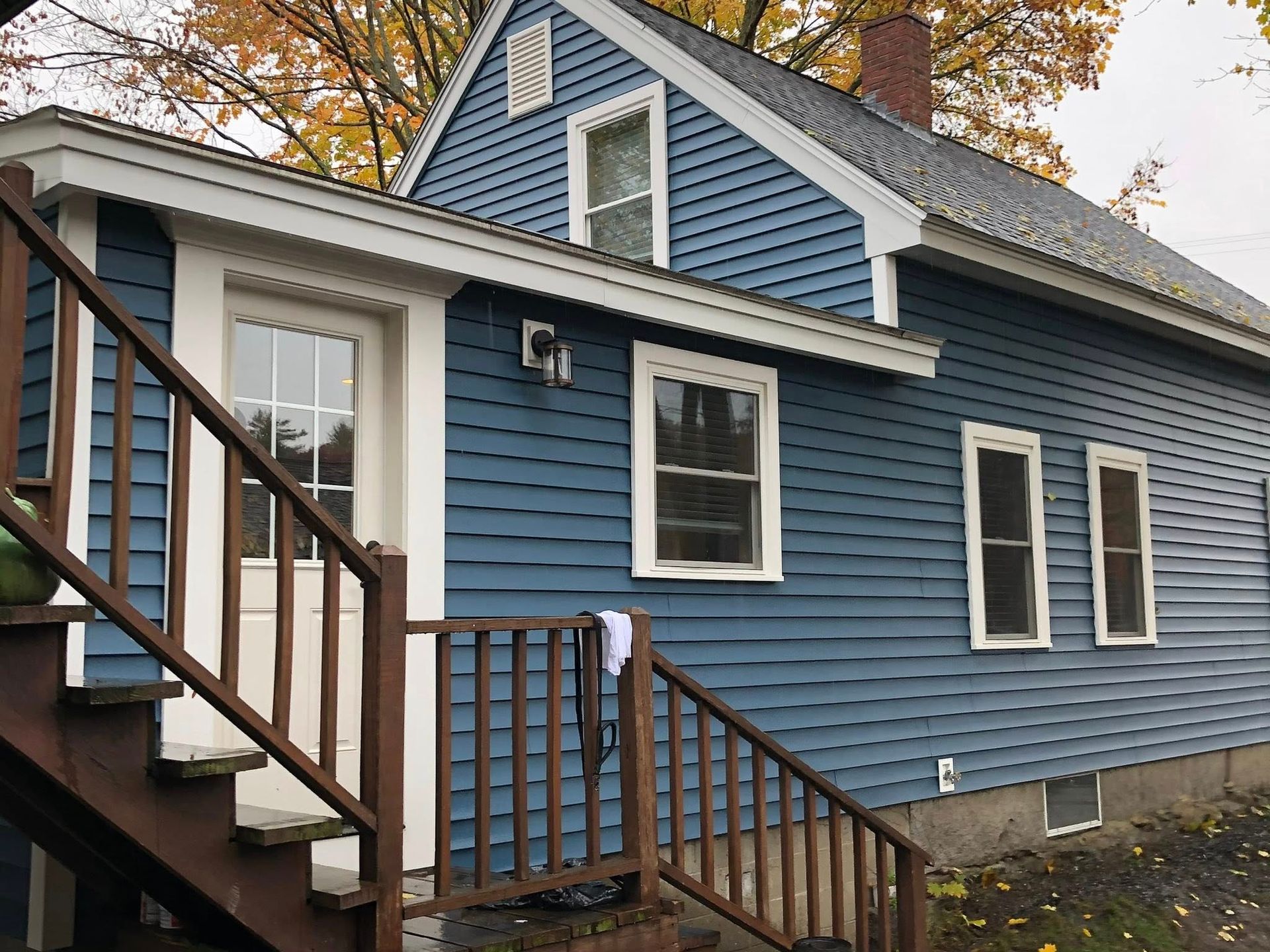 A blue house with white trim features a wooden staircase leading to a side door and several windows on a cloudy day.