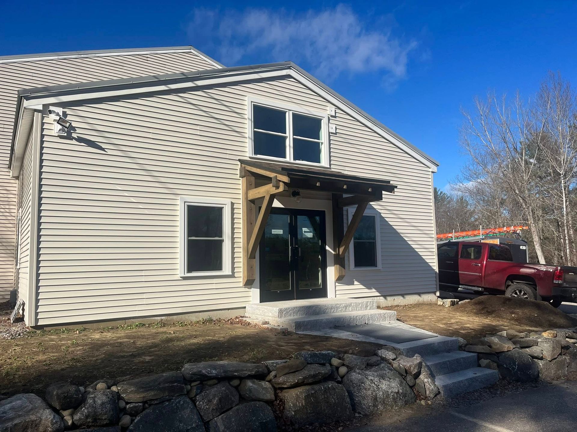 A two-story building with beige horizontal siding, a wooden front portico, and a stone retaining wall in the foreground.