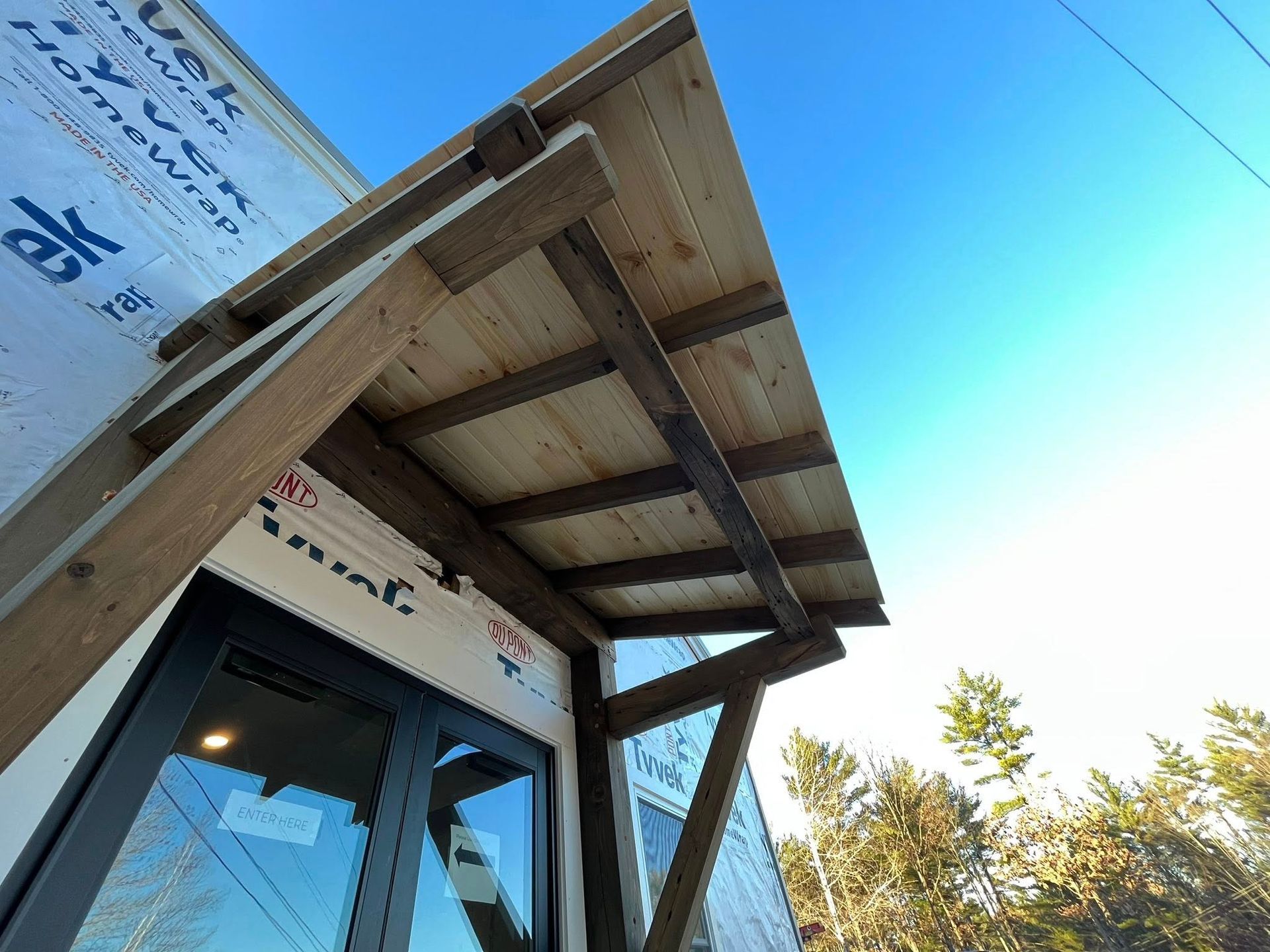Low-angle view of a wooden porch roof canopy being built over a glass door on an unfinished building exterior.