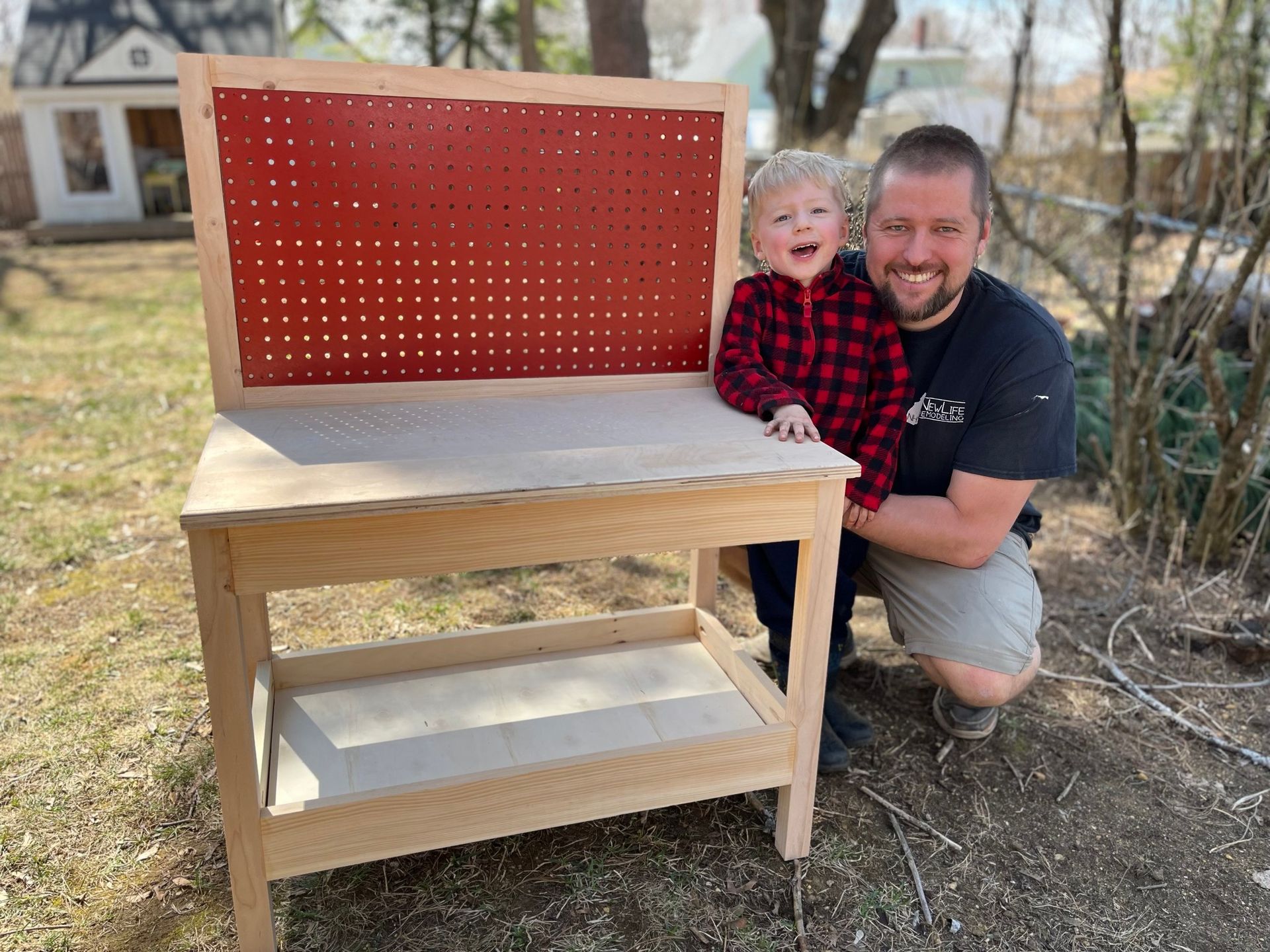 A man and a child smile next to a newly built wooden workbench with a red pegboard, outdoors in a yard.