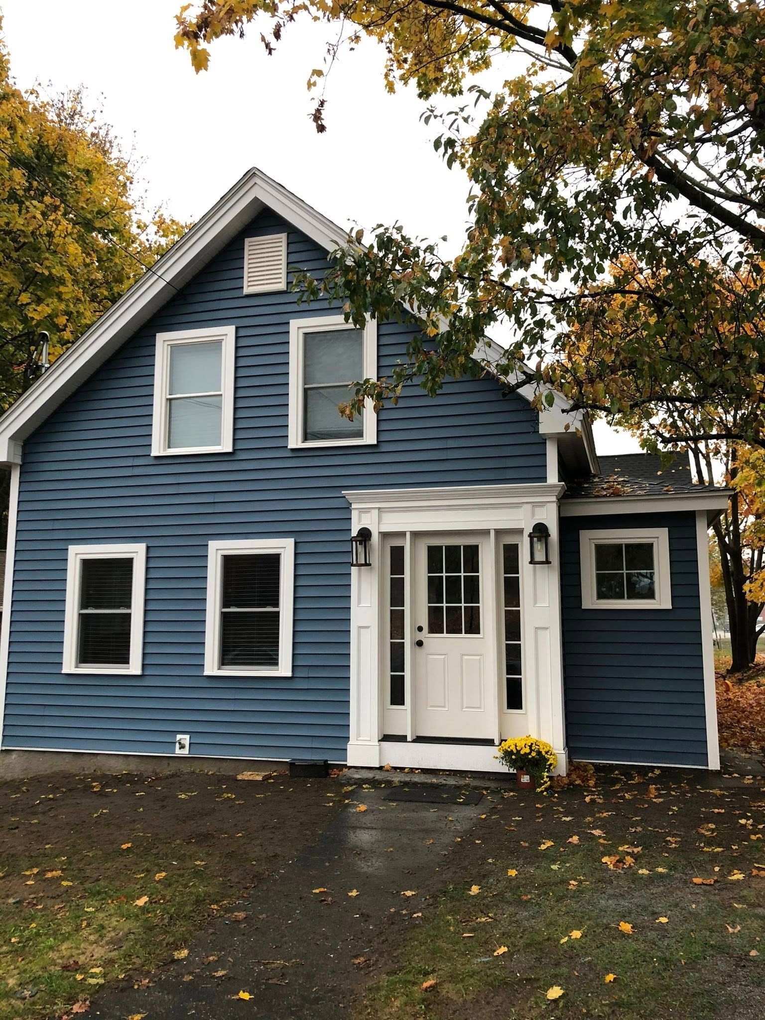 A two-story blue house with white trim and a small front porch, surrounded by trees with autumn leaves.
