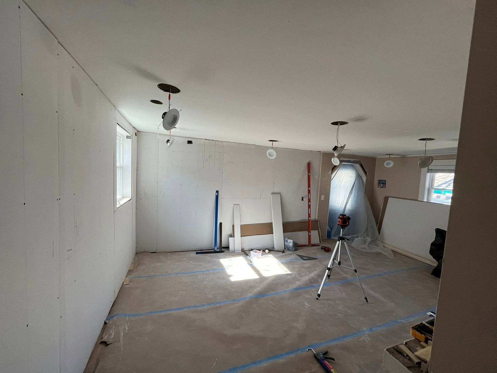 A room under construction with white drywall on walls and ceiling, a laser level on a tripod, and tools on the floor.