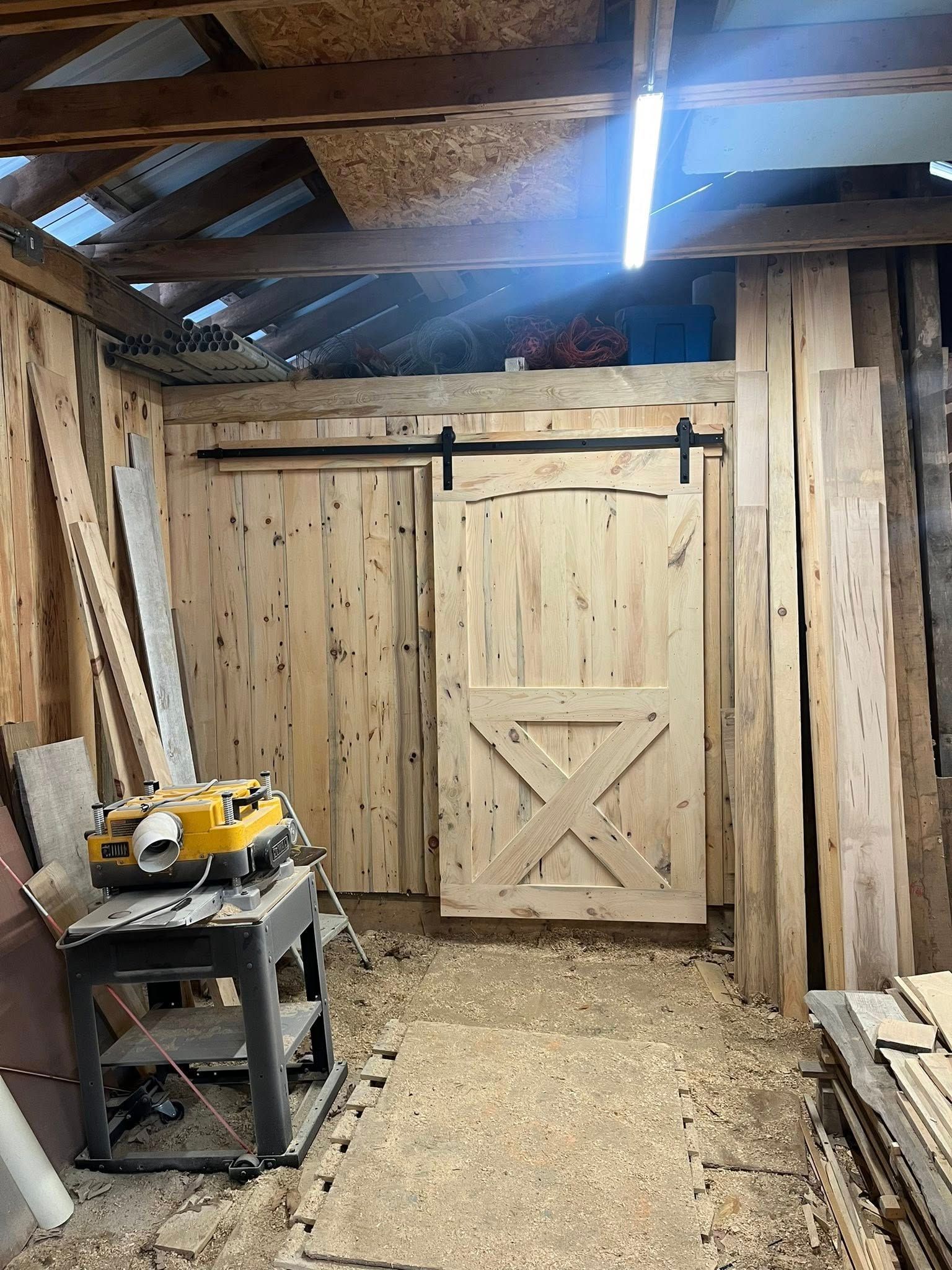 A sliding barn-style wooden door in a rustic workshop filled with stacked lumber and a woodworking planer on a stand.