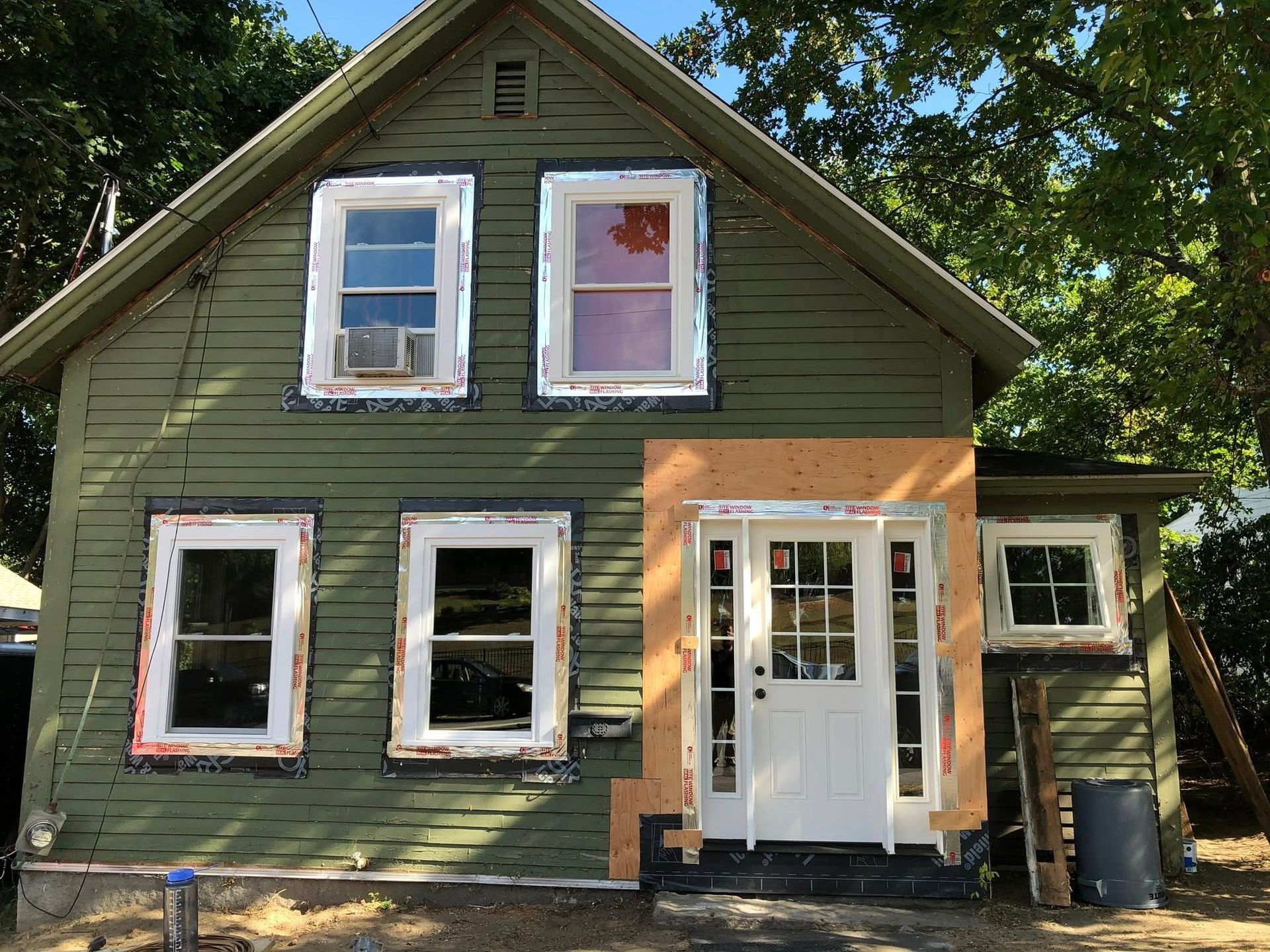 An olive-green house under renovation with a newly installed white front door and taped window frames.