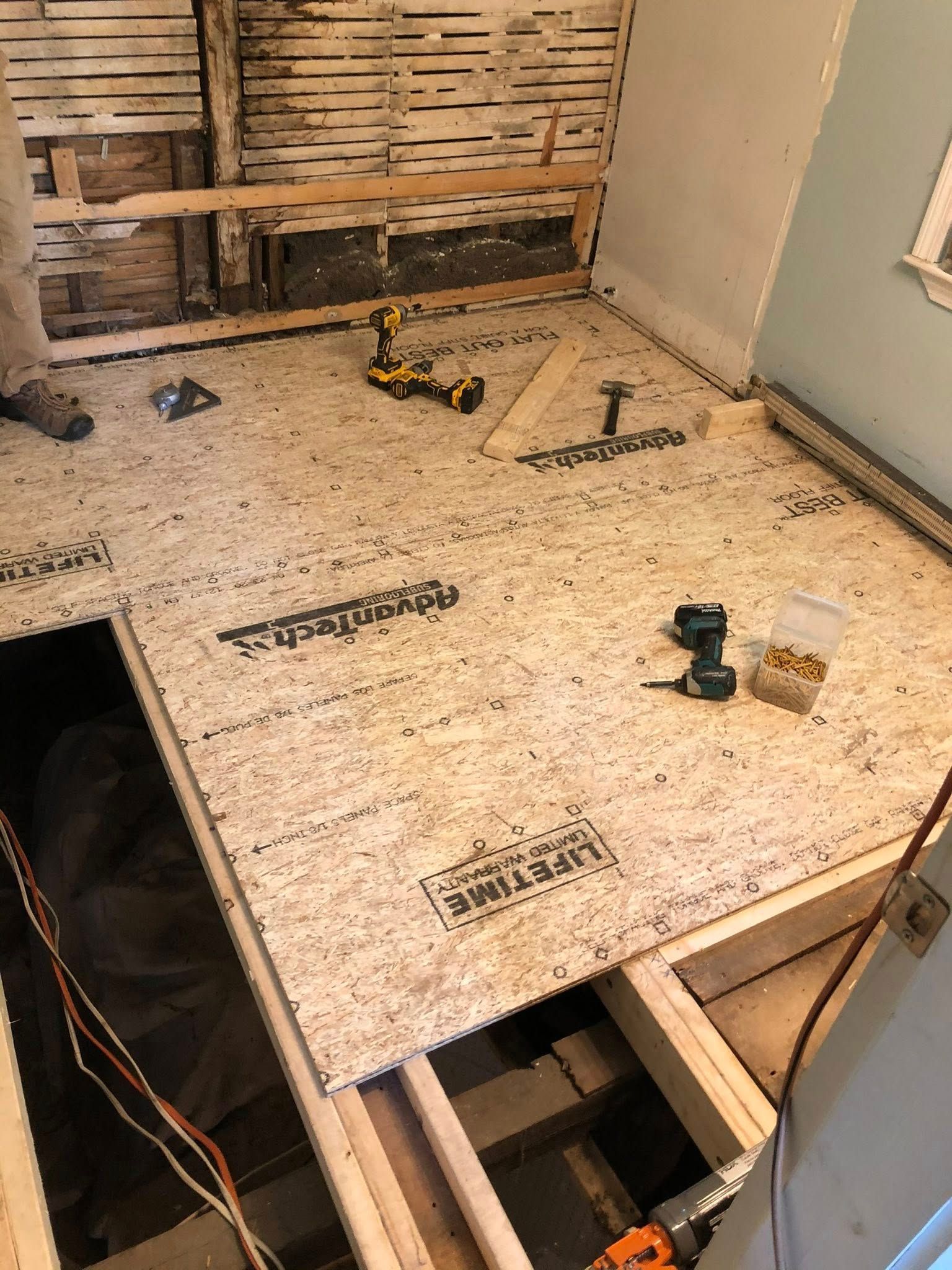 A room under renovation with partially installed subflooring, exposed wooden studs, and tools scattered on the floor.