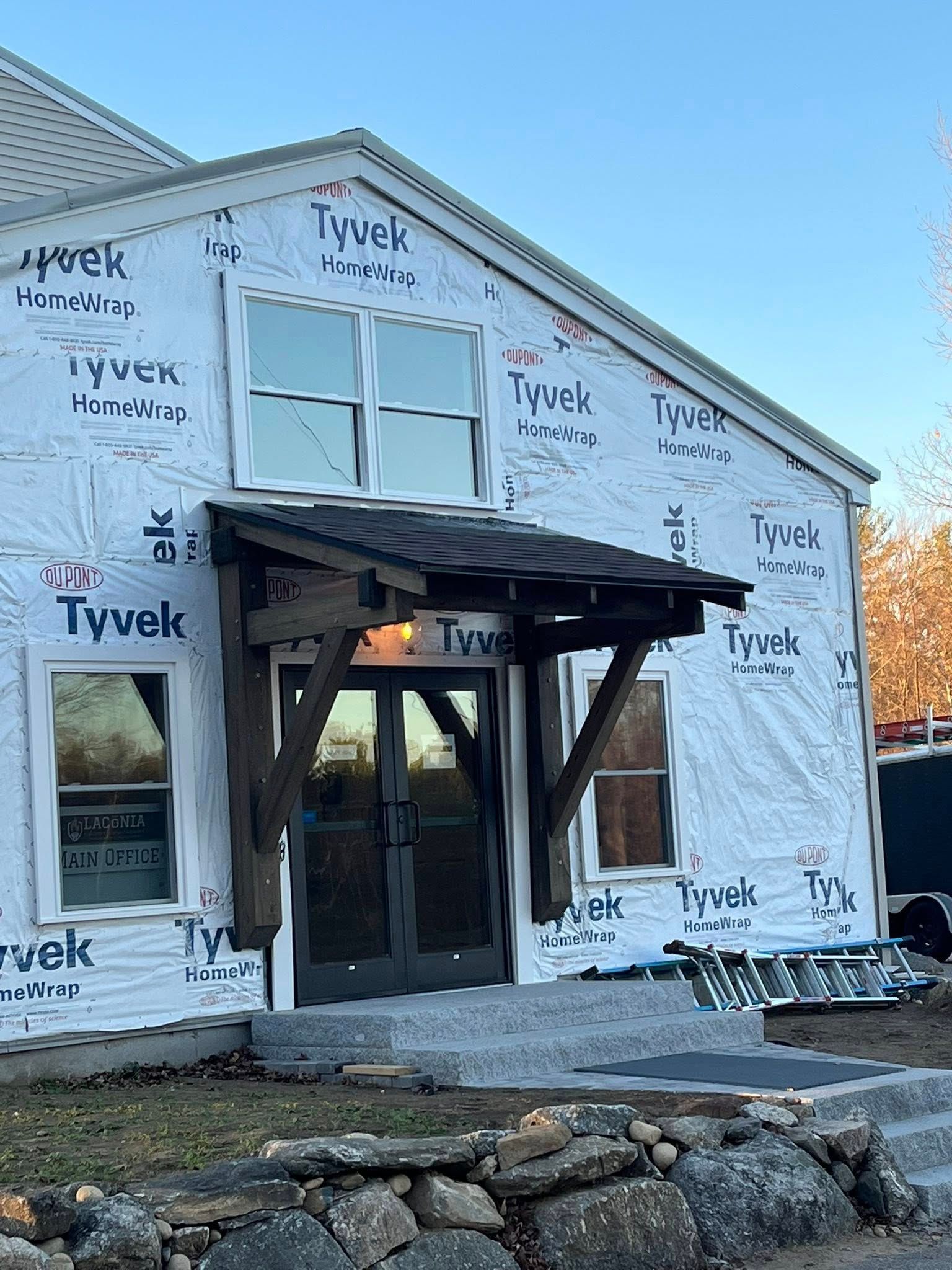 Exterior of a building under construction, featuring white Tyvek wrap, dark double doors, a timber porch, and stone steps.