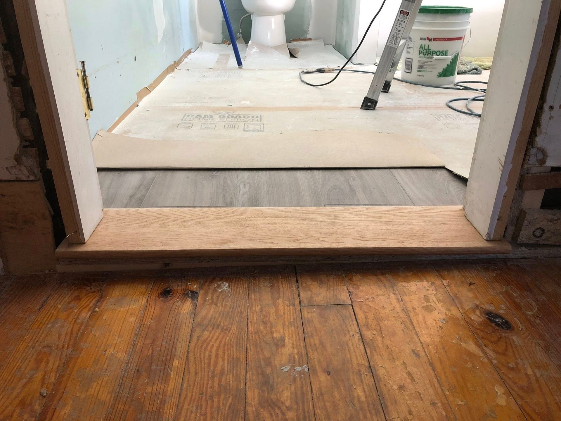 A doorway threshold transitioning from hardwood flooring to a bathroom under renovation with new gray flooring and a toilet.