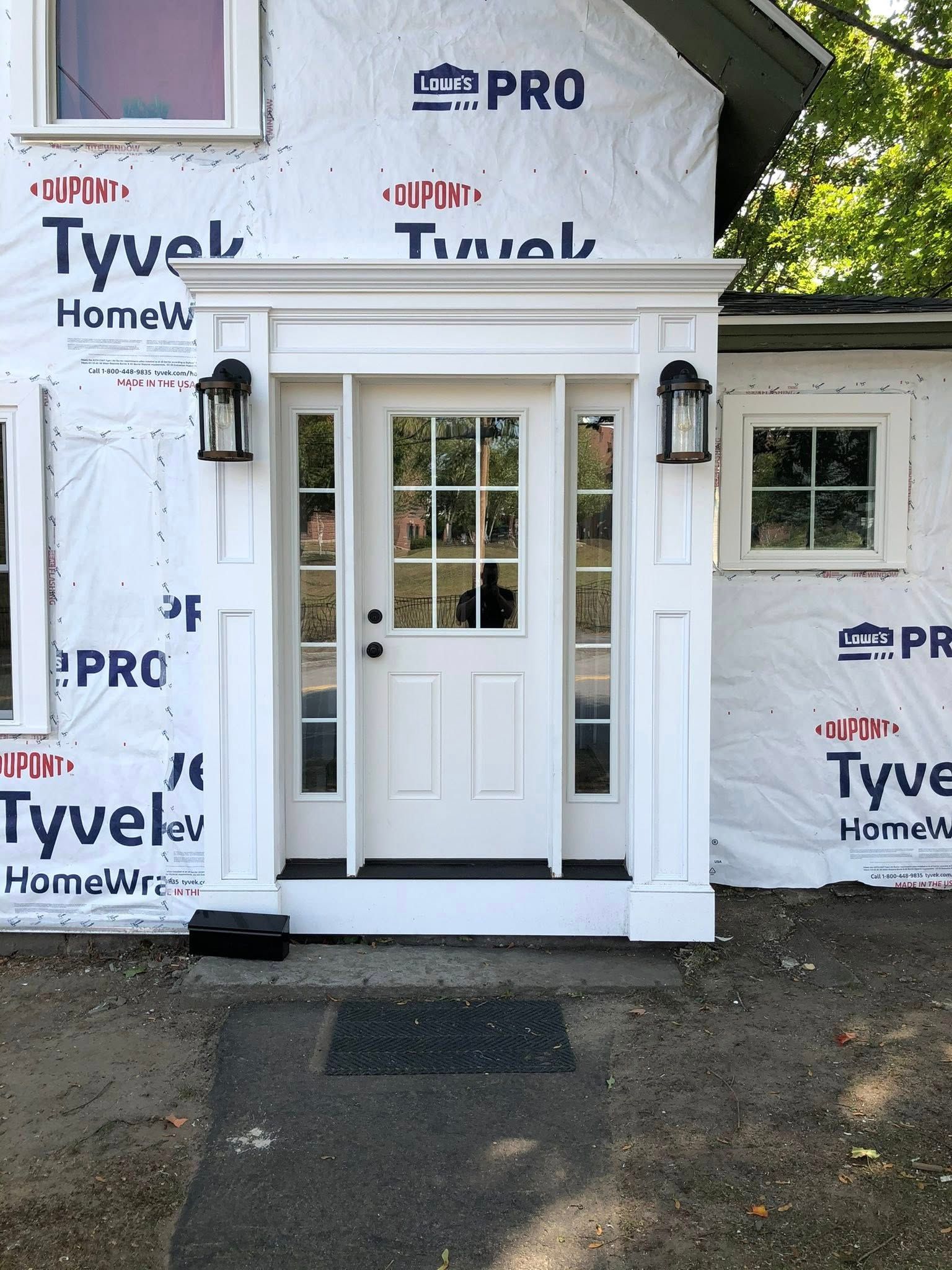 A white entry door with side windows and a decorative surround installed in an unfinished house exterior with Tyvek wrap.