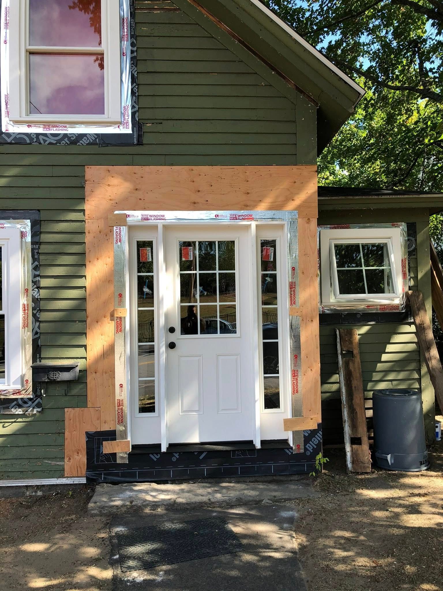 A newly installed white front door with sidelights surrounded by plywood and flashing on an olive-green wooden house.