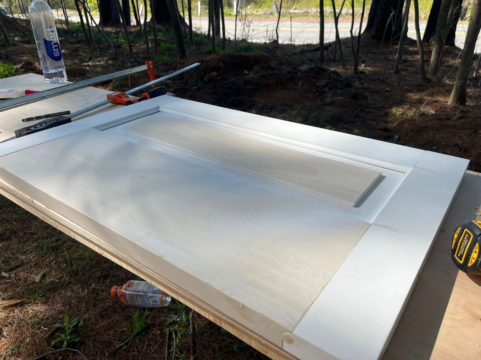 A white cabinet door panel rests on a workbench outdoors, secured by a metal clamp, with wood panels visible nearby.