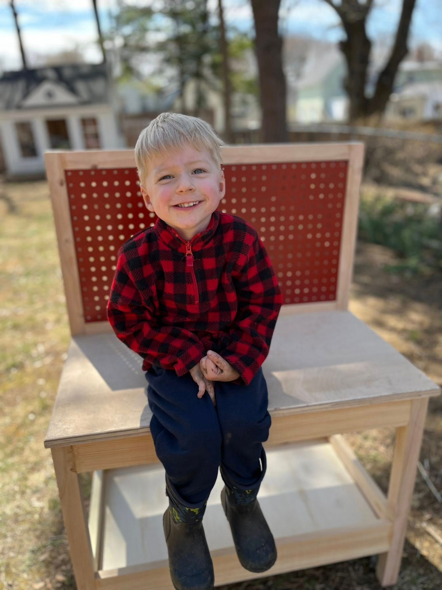 A young person in a red and black plaid shirt and blue pants smiles while sitting on a wooden bench outdoors.