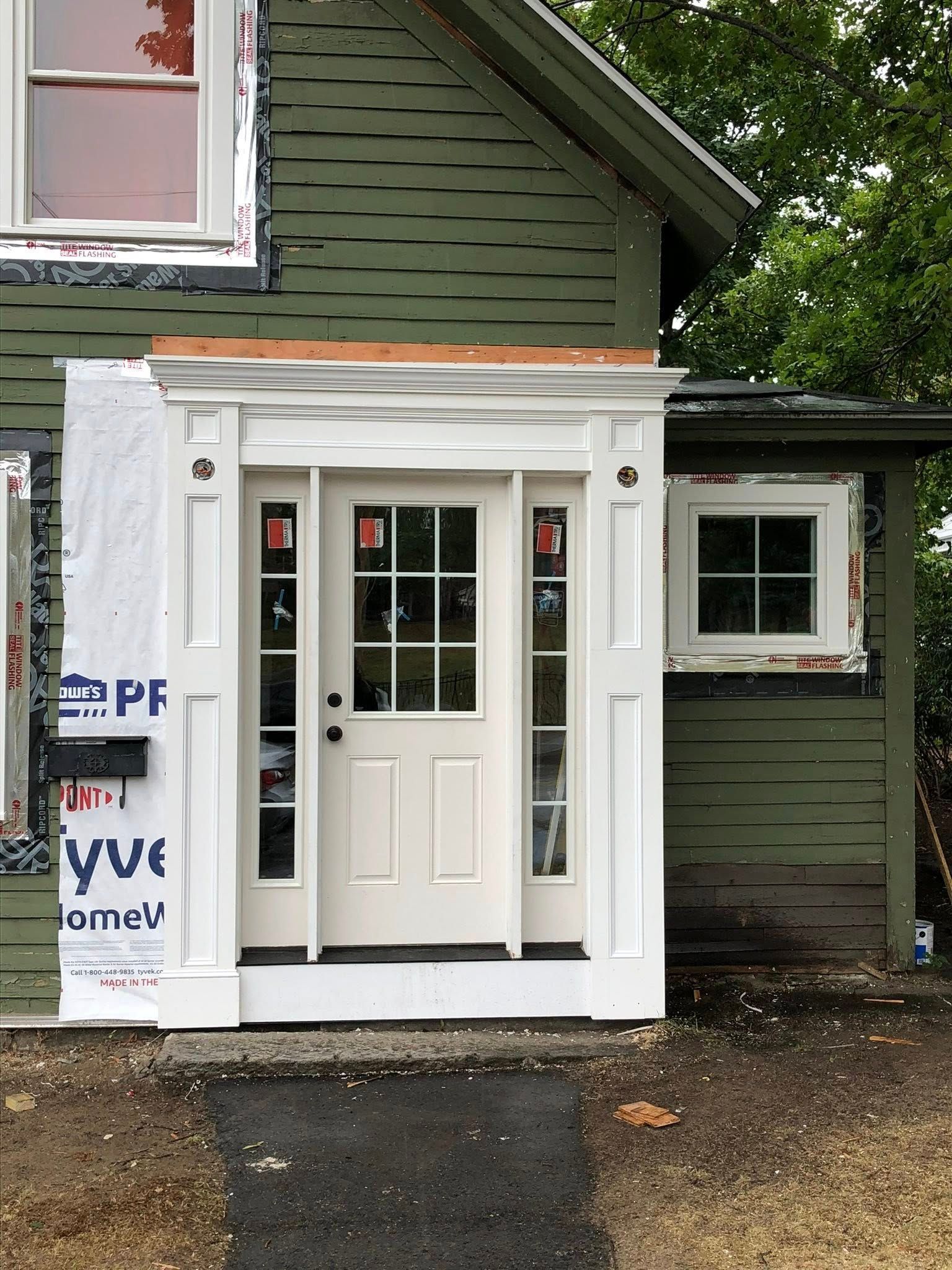 A newly installed white front door with side panels, framed in white trim, on the side of a green wood-sided house.