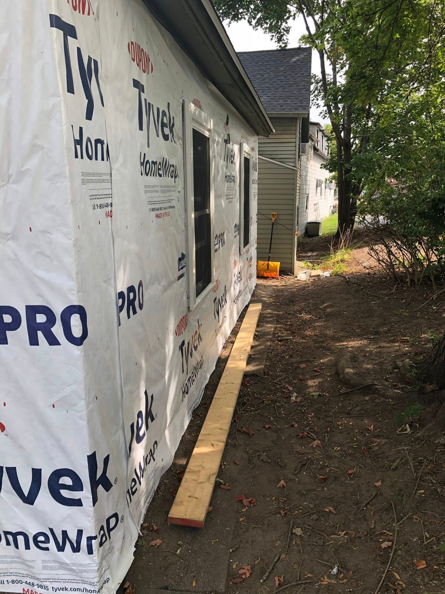 A house exterior wrapped in white Tyvek, with a wooden board on the dirt ground beside it near another house and trees.