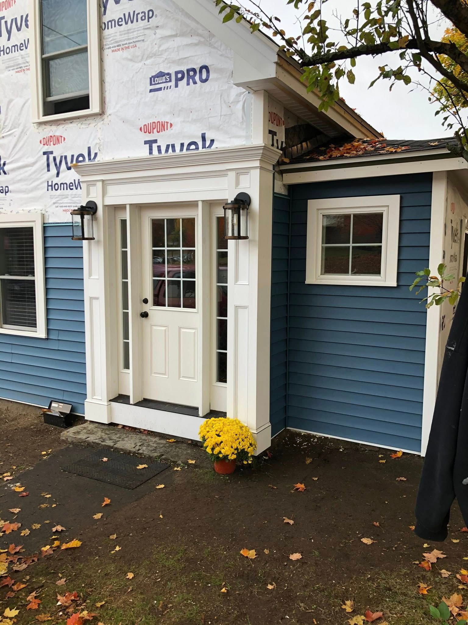 Exterior view of a blue house with white trim, a central front door, and ongoing construction with exposed Tyvek wrap.