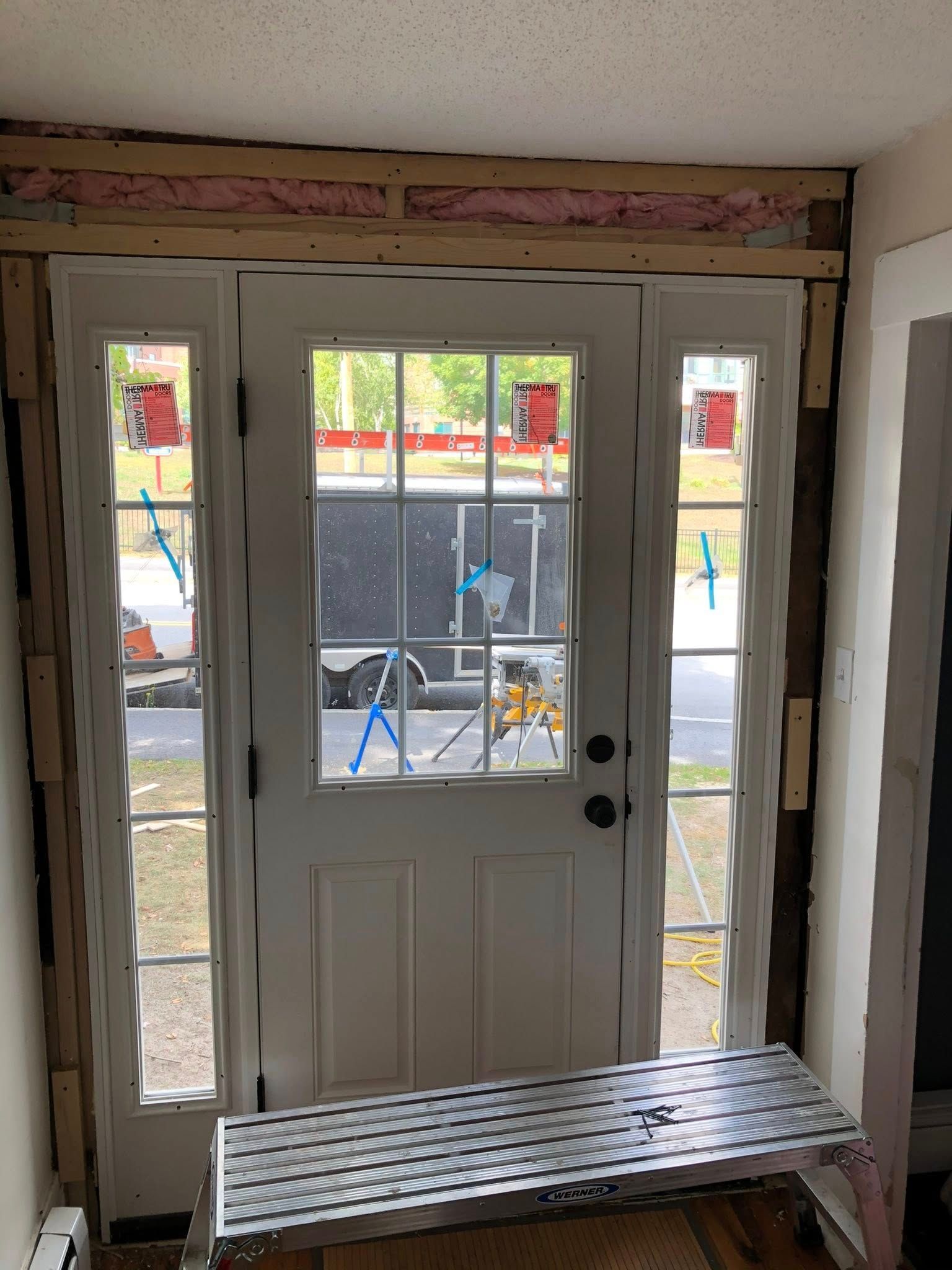 A newly installed white front door with sidelights surrounded by exposed framing, insulation, and a metal step platform.
