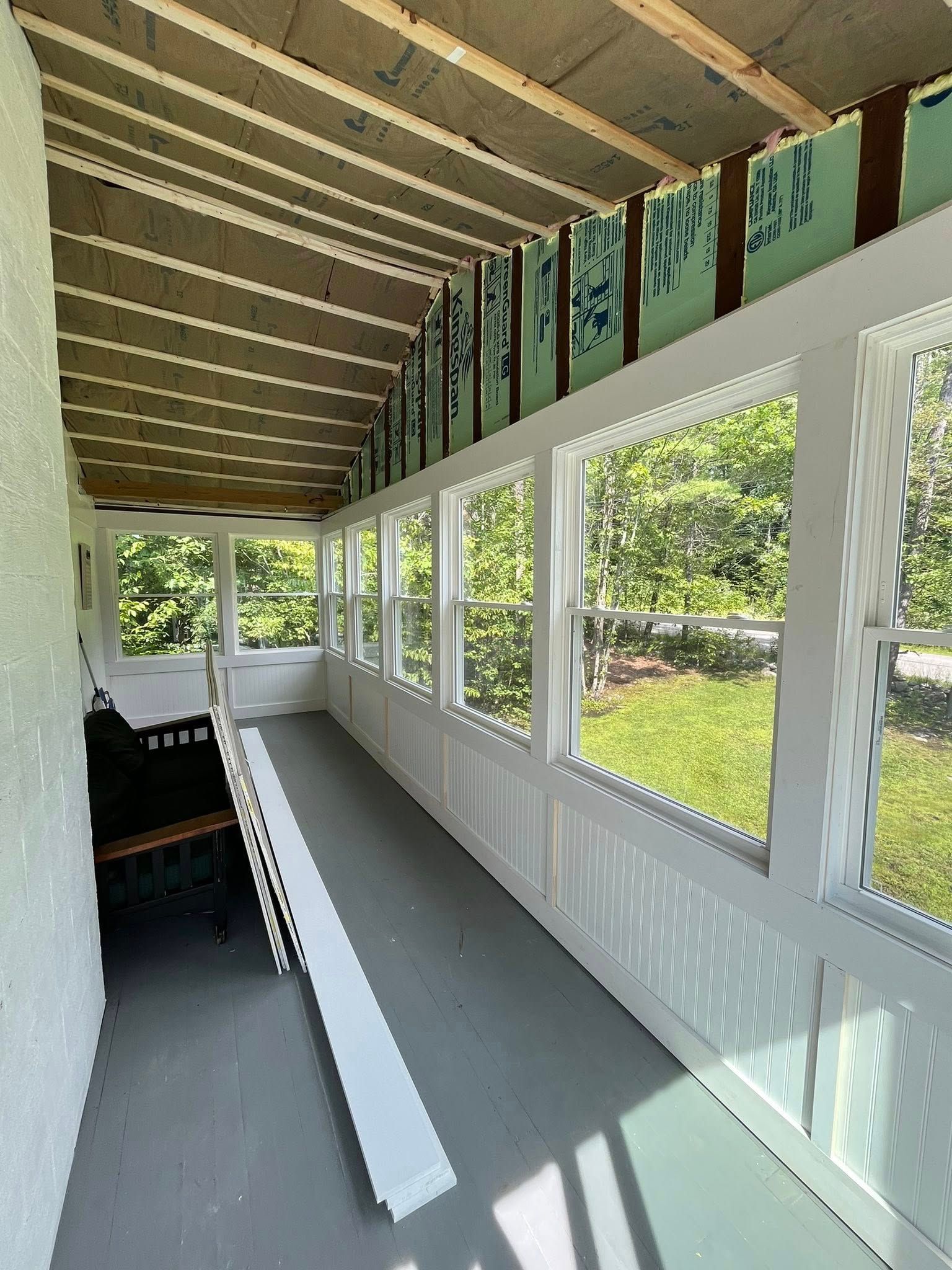 An unfinished enclosed porch with white framed windows, exposed ceiling beams, and a piece of white trim leaning on the floor.