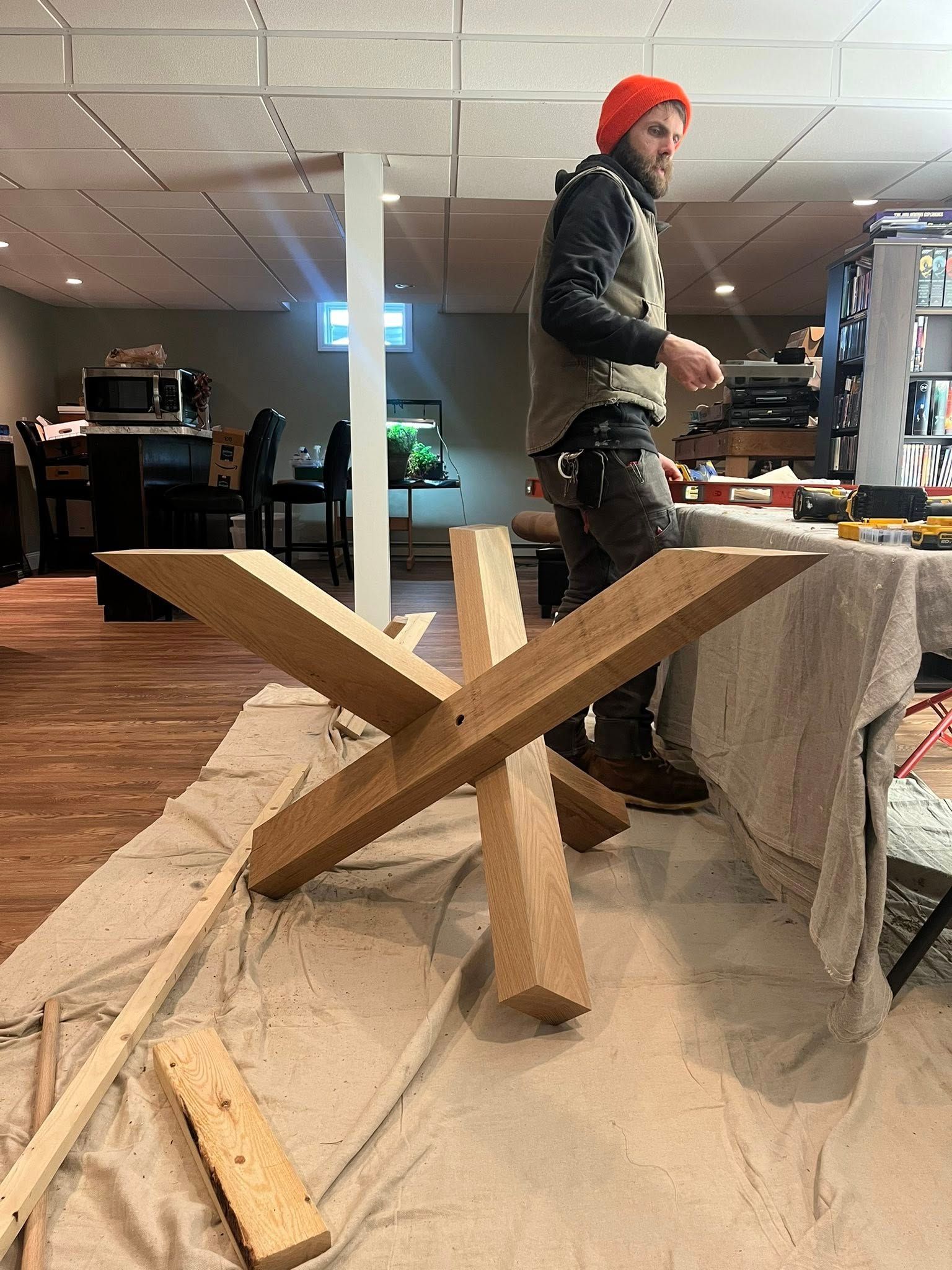 A person in a red beanie and work vest assembles a large, X-shaped wooden table base in a room.