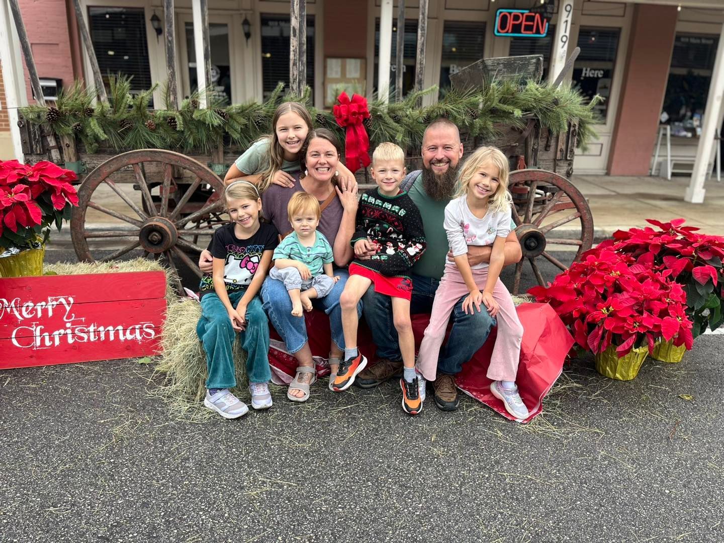 A family smiles together for a Christmas photo, posing in front of a wooden wagon decorated with greenery and poinsettias.