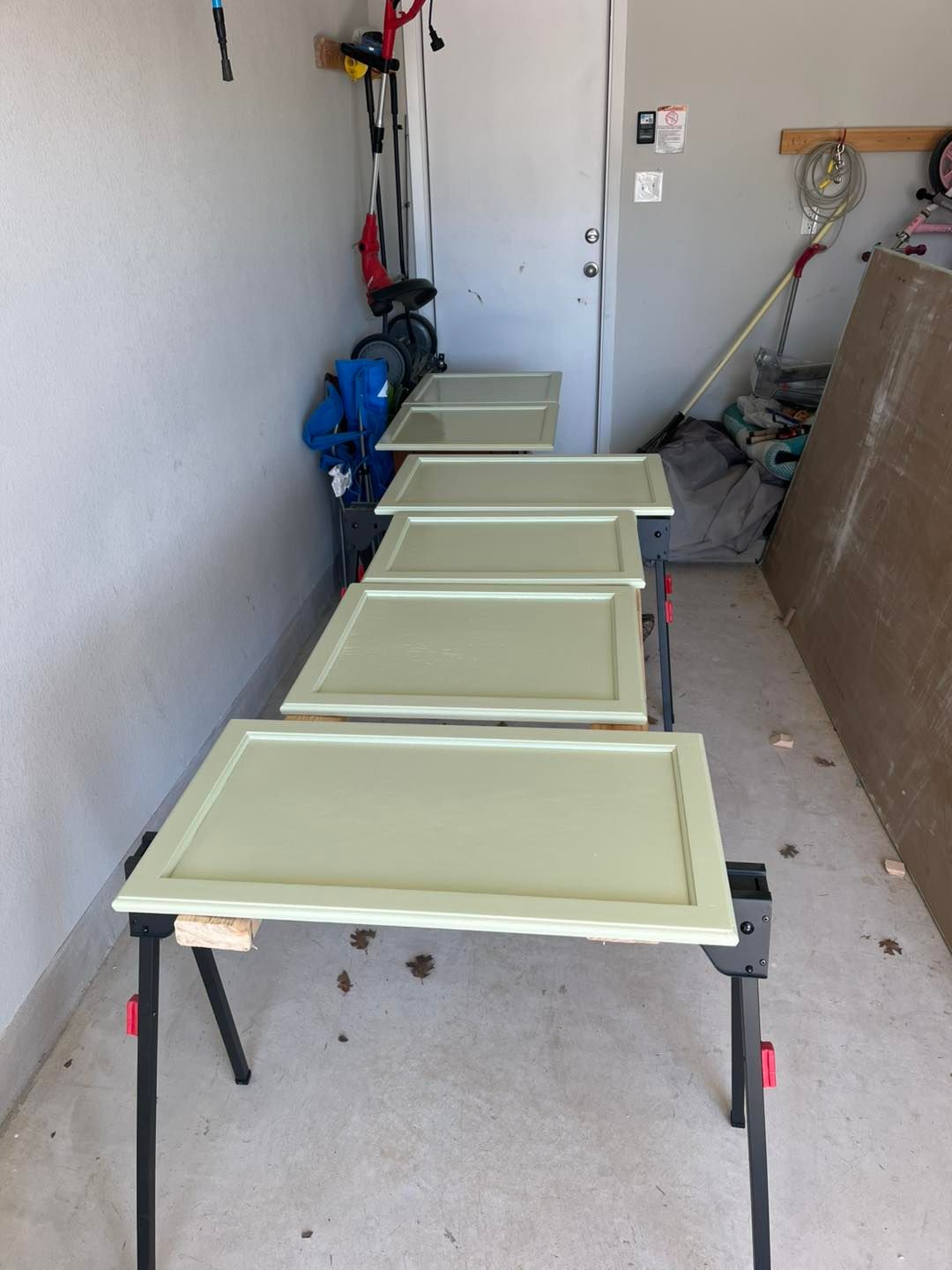 Five pale green, rectangular cabinet doors are lined up on a workbench in a garage setting.
