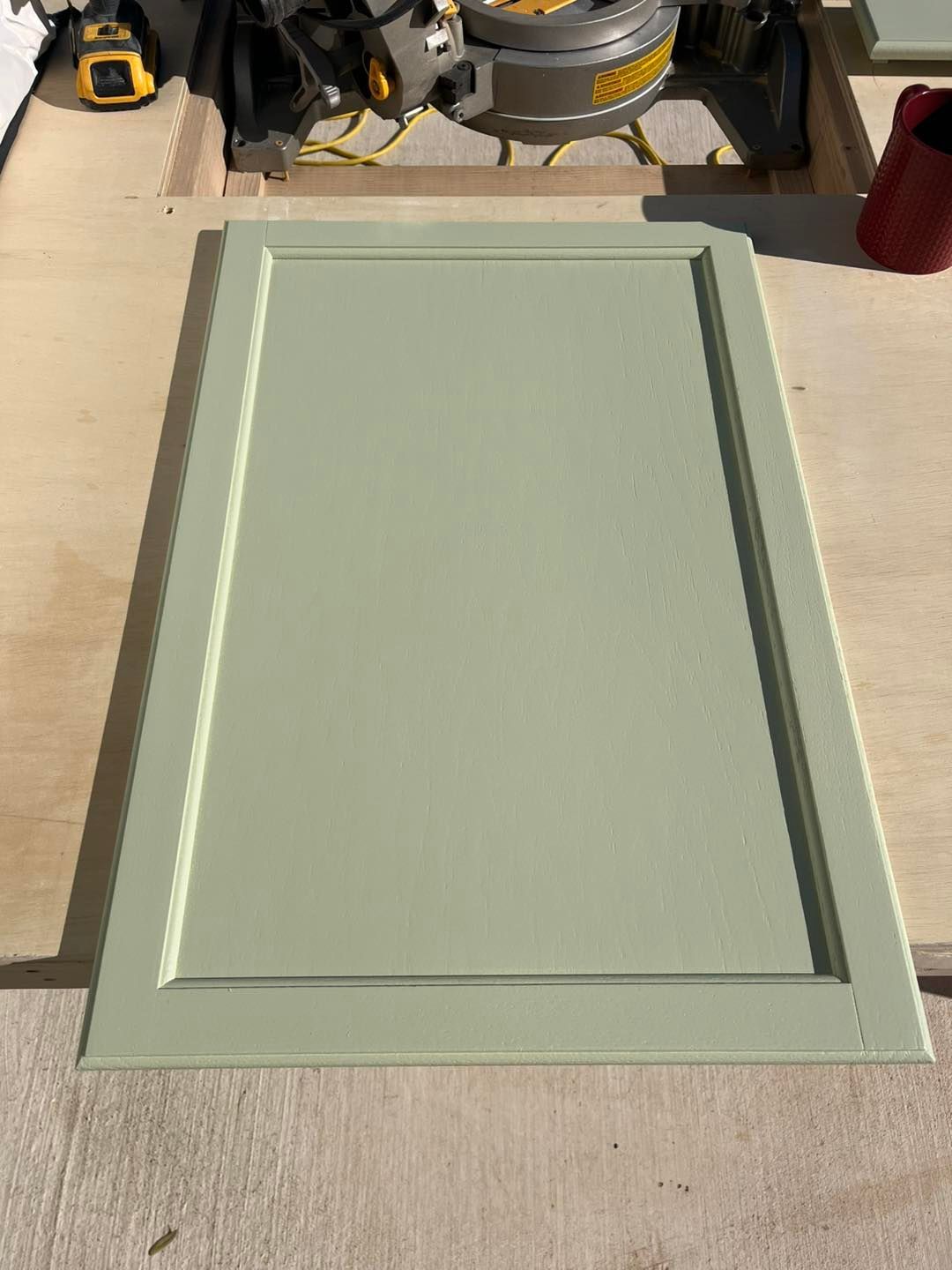 A sage green, rectangular cabinet door with a recessed center panel sits on a light-colored wood workbench.