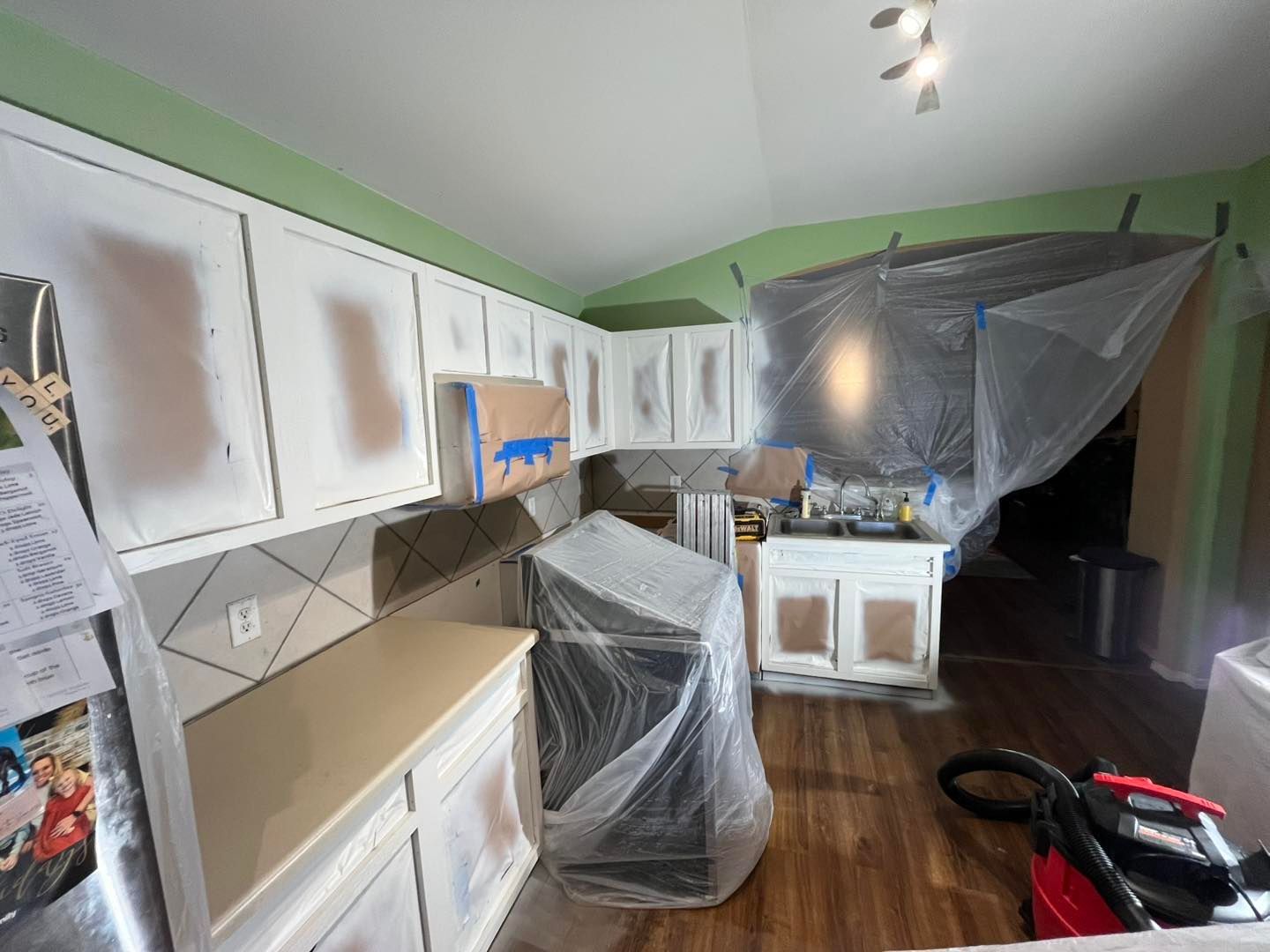 Kitchen undergoing cabinet painting, with cabinets and appliances covered in plastic sheeting and tape.