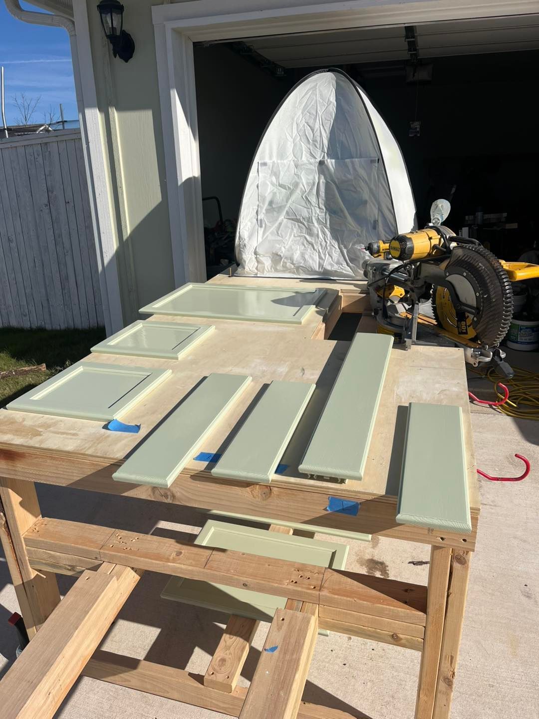 Freshly painted sage green cabinet doors drying on a wooden workbench in a garage with a miter saw in the background.