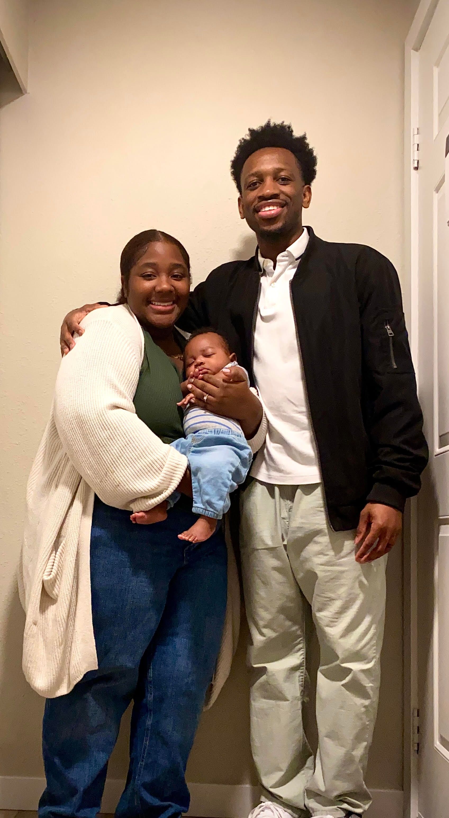 Family of three smiling, posing indoors: person holding infant, other person has arm around the holding person.
