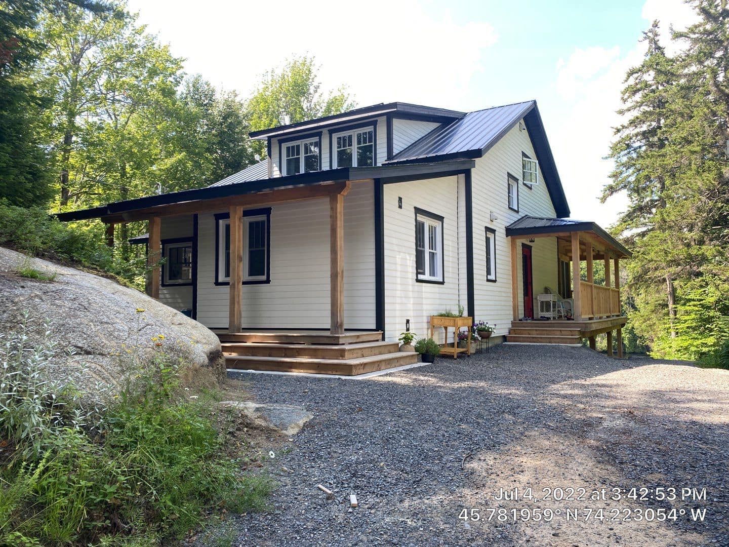A white house with a black roof and a porch is surrounded by trees.