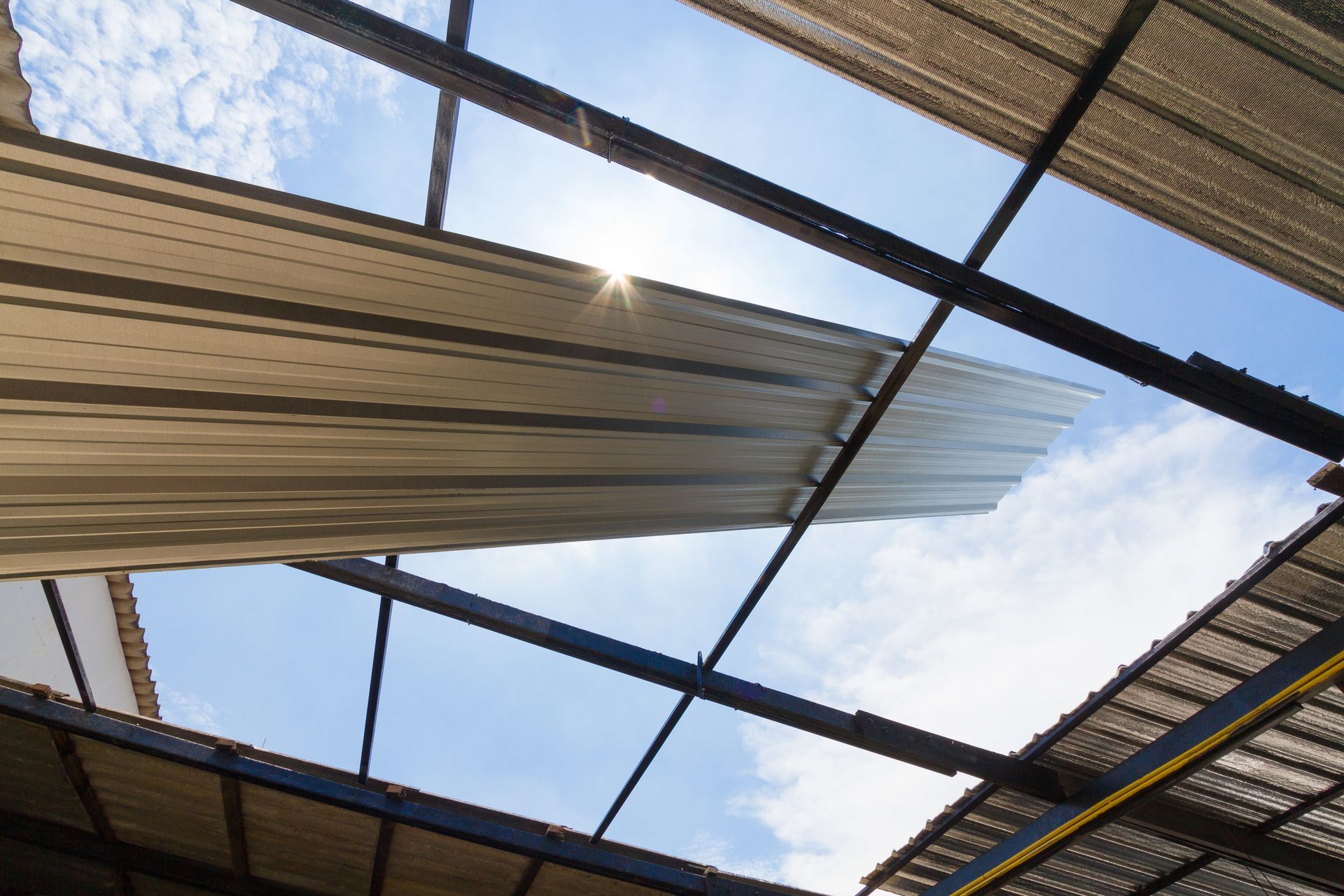 Looking up at a partially collapsed roof with metal panels against a blue sky with sunlight.