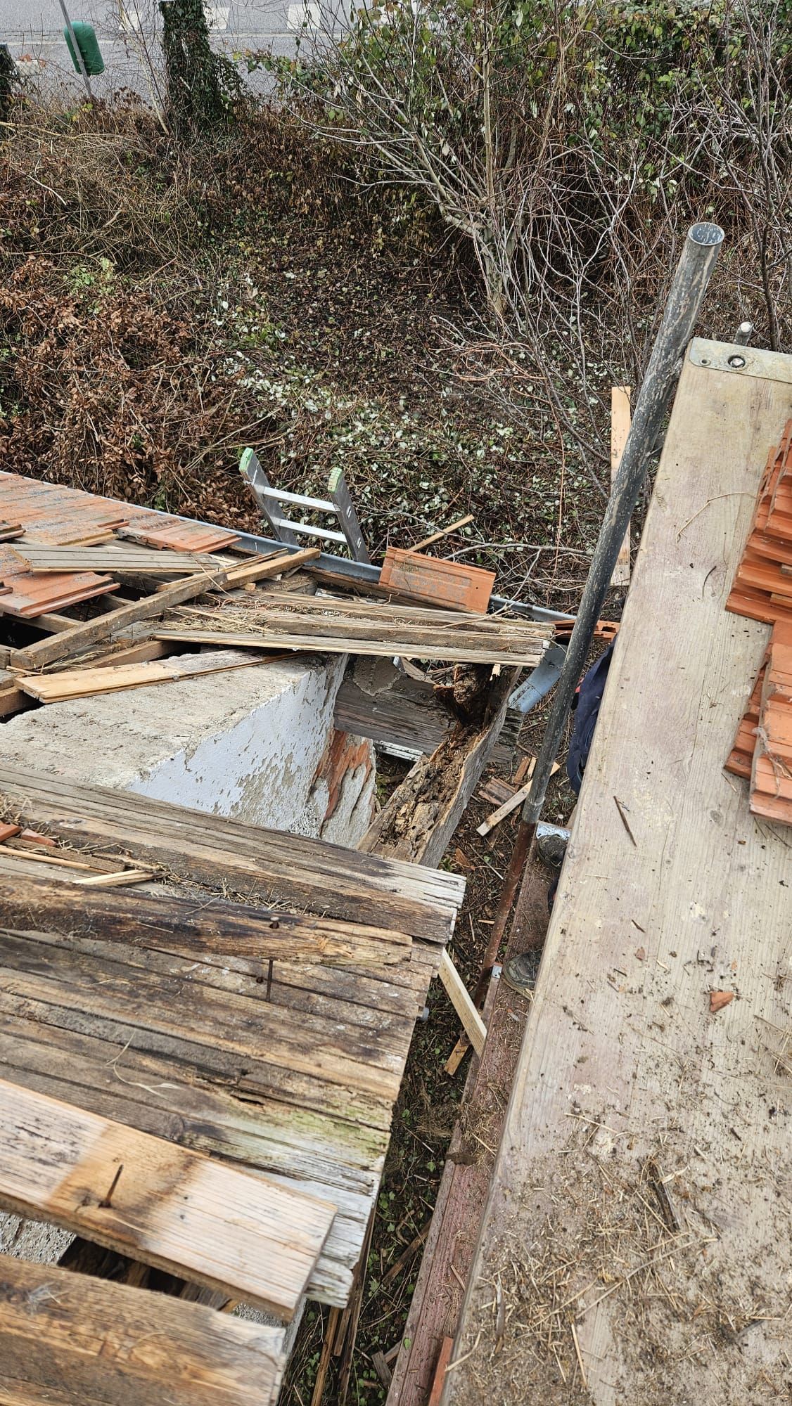 Beschädigtes Dach mit zerbrochenem Holz, Ziegeln und einem langen, dunklen Gegenstand, der an einem Betonteil lehnt; Außenaufnahme.