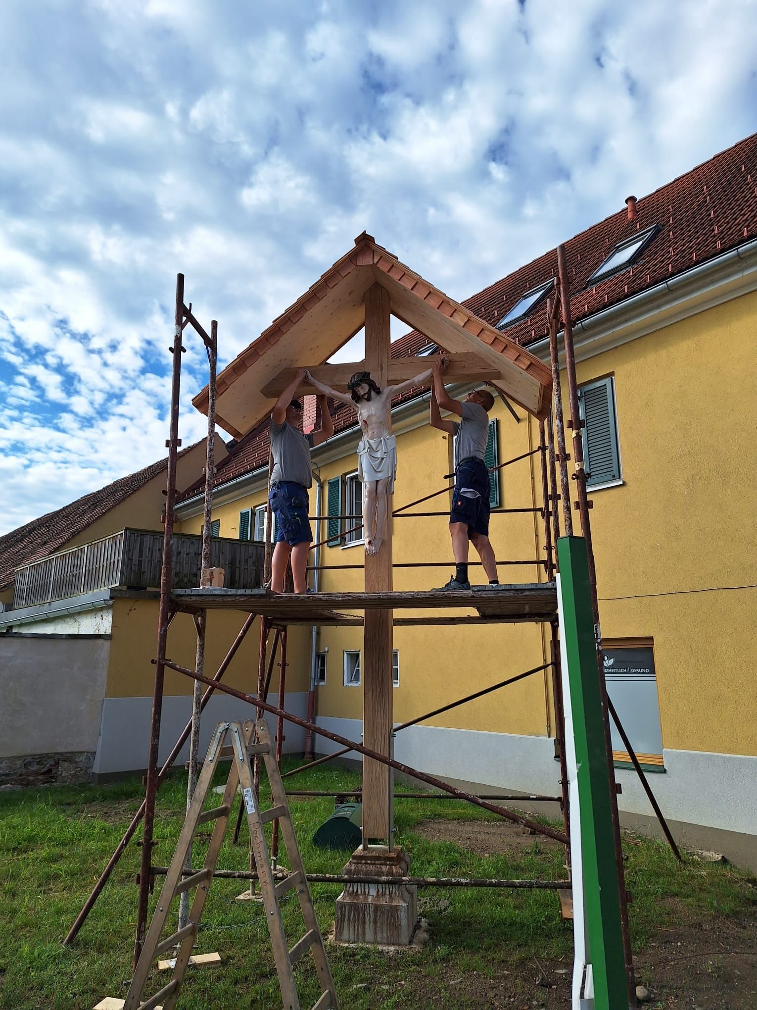 Zwei Männer arbeiten auf einem Gerüst vor einem gelben Gebäude an einer Holzkonstruktion.