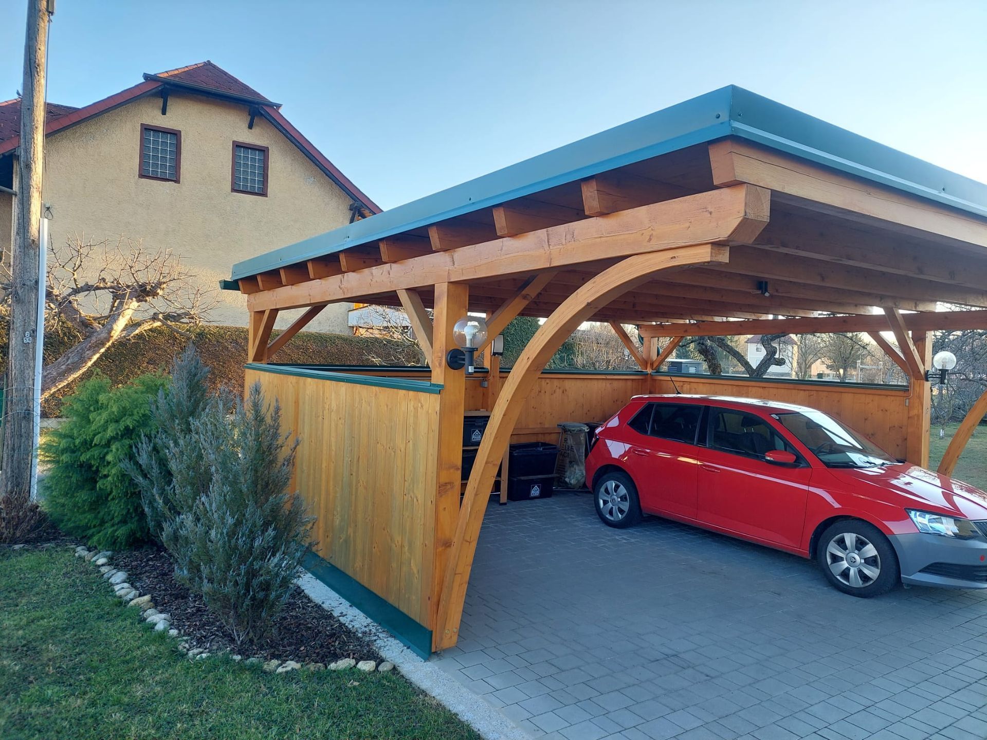 Ein rotes Auto ist unter einem hölzernen Carport geparkt.